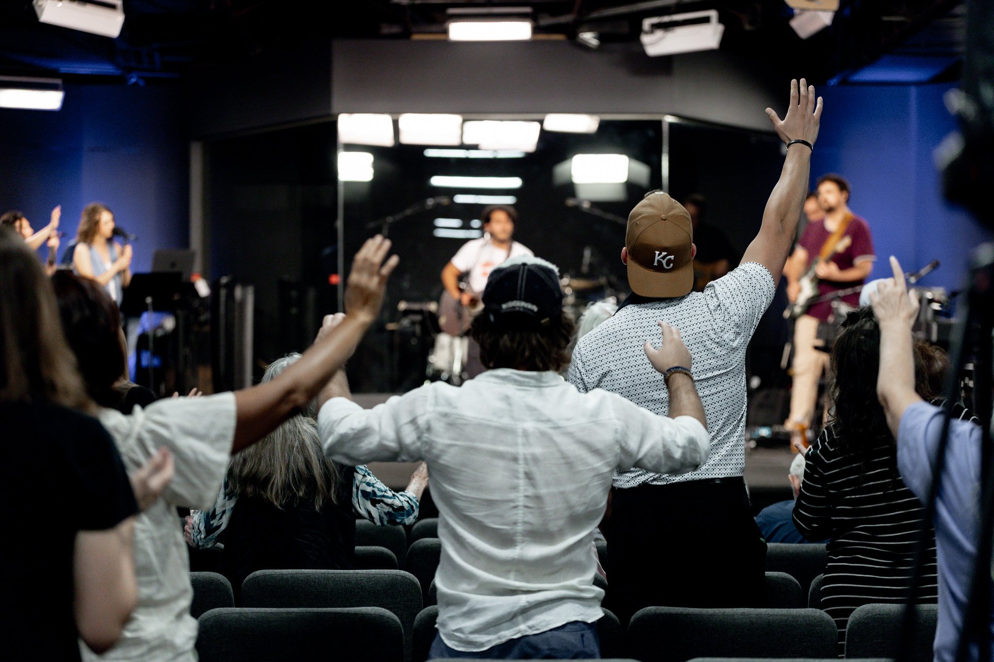 A group of people in a crowded indoor setting, raising their hands, with a person at the front playing guitar and others singing or speaking into microphones.