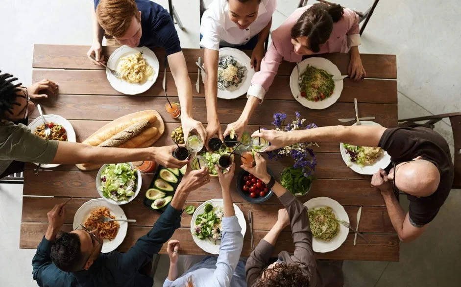 Top-down view of a diverse group of people raising glasses of drinks over a wooden dining table filled with various salads, bread, cherry tomatoes, and a vase with purple flowers, celebrating together.