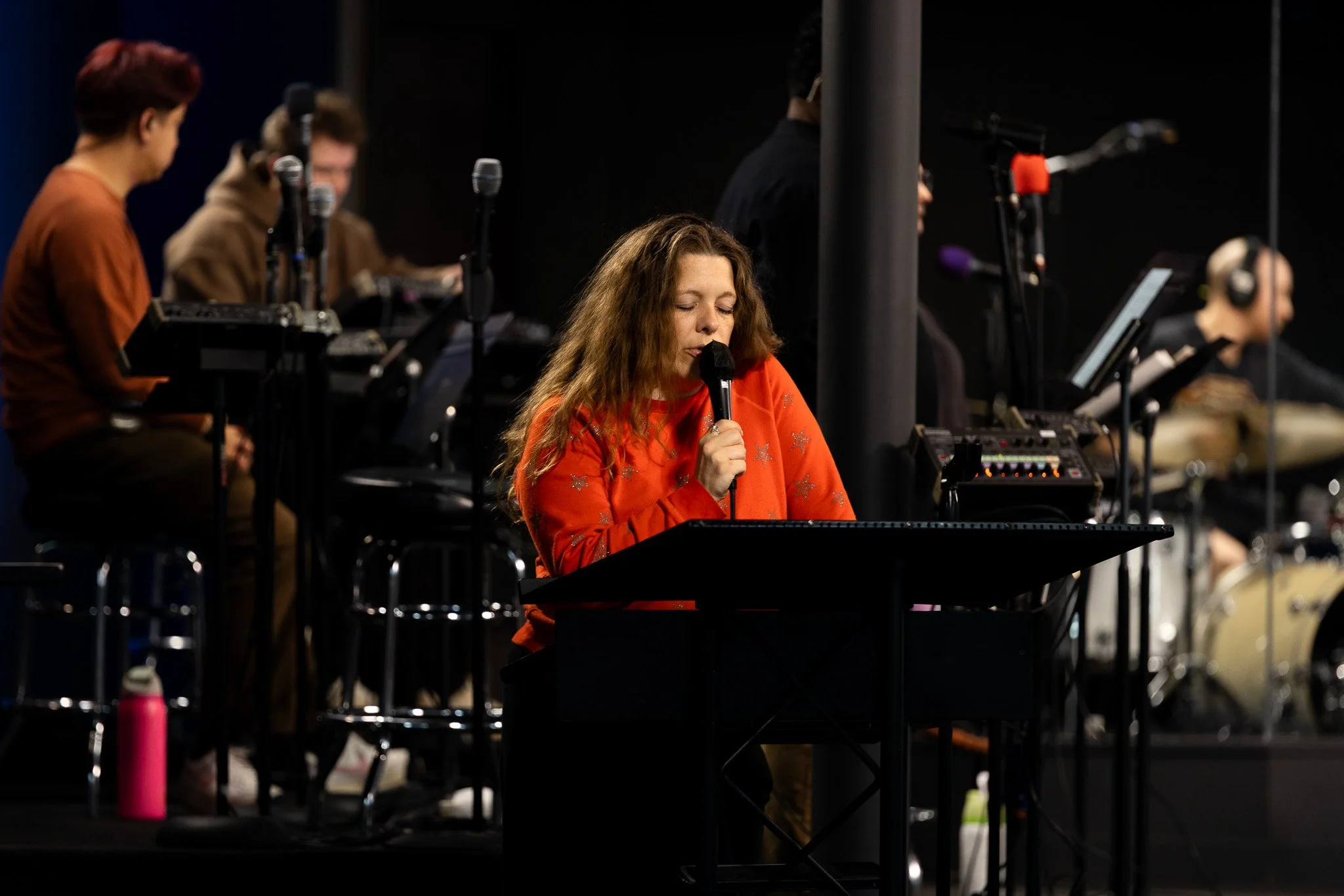 A woman with curly hair wearing a red top with silver star patterns, singing into a microphone while playing a keyboard in a dark setting with other musicians in the background.
