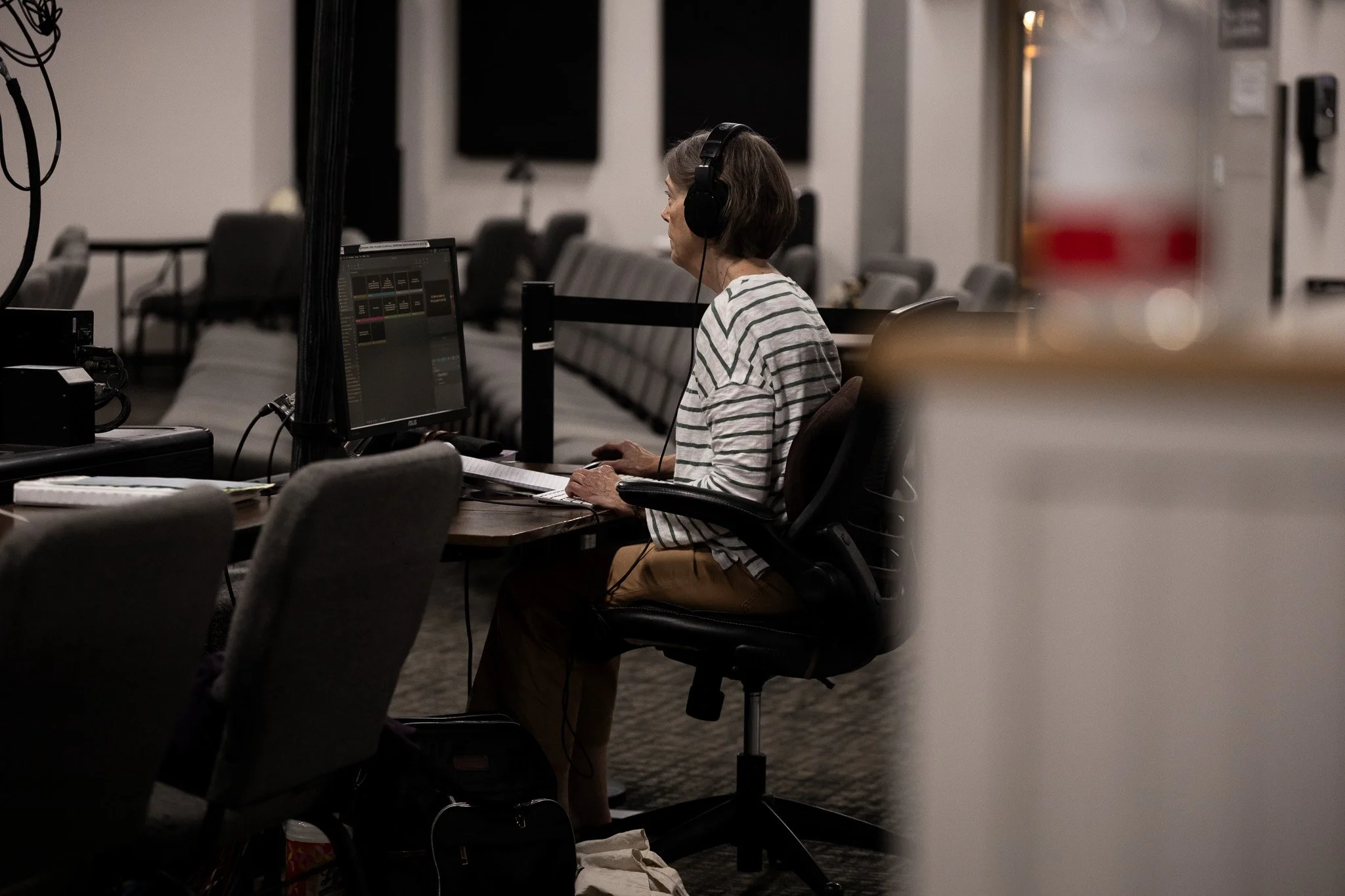 A woman with brown hair wearing a striped shirt and tan pants, sitting at a desk with a computer and wearing black headphones, in a room with rows of chairs and dark wall-mounted screens.
