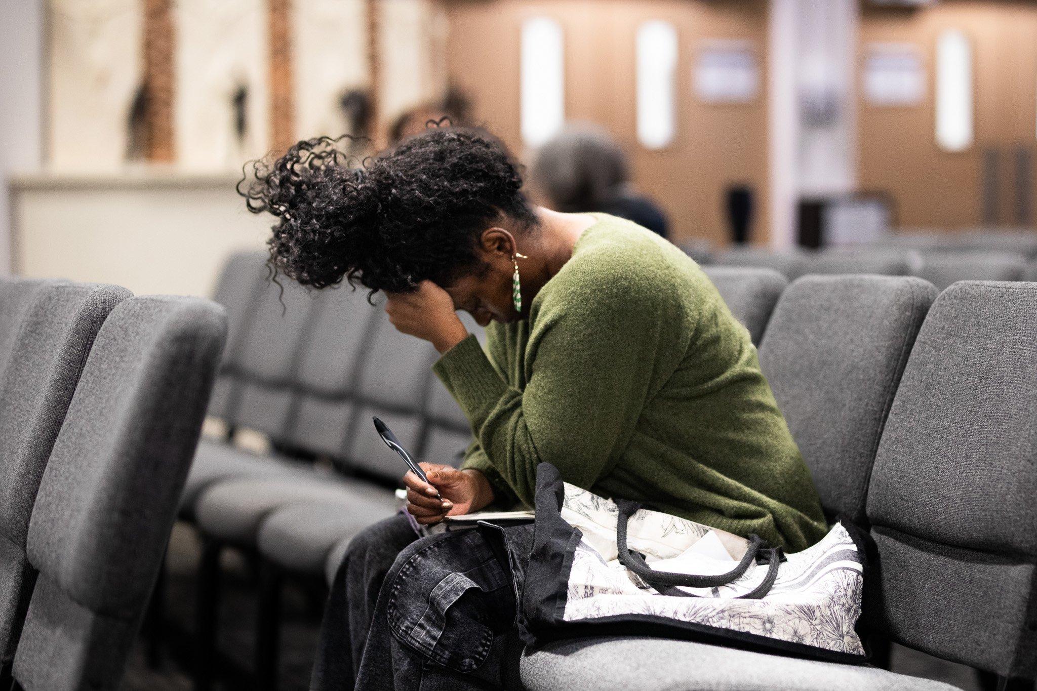 A woman with curly hair wearing a green sweater sitting in an auditorium, looking down at her phone with a pen in her hand, with other people seated behind her.