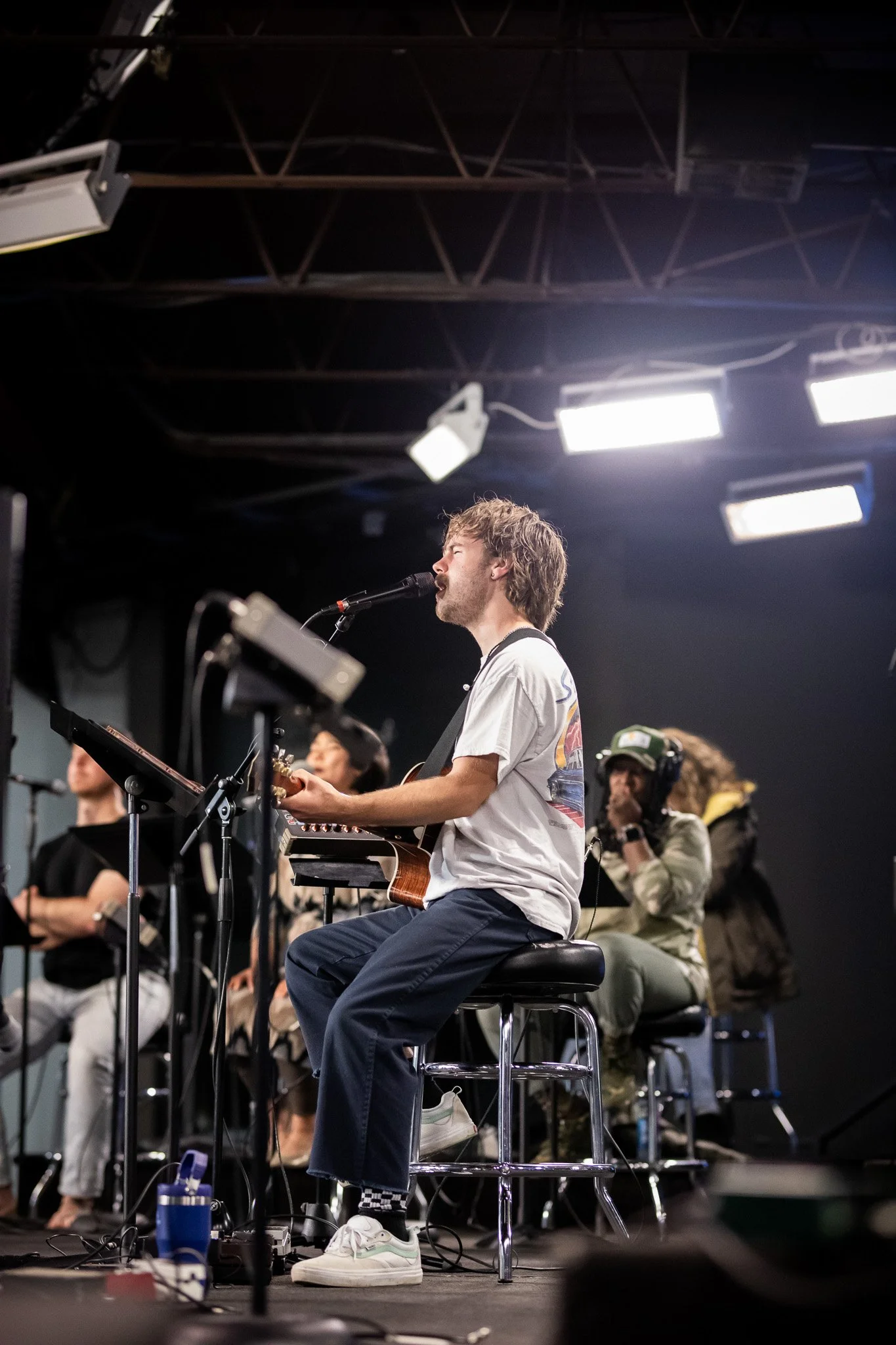 A person with curly hair singing into a microphone, seated on a stool, playing an acoustic guitar on stage during a performance with an audience in the background.