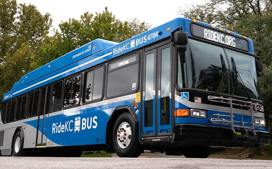 A blue and white RideKC Bus parked outdoors with trees in the background, displaying the website RideKC.org on the front.
