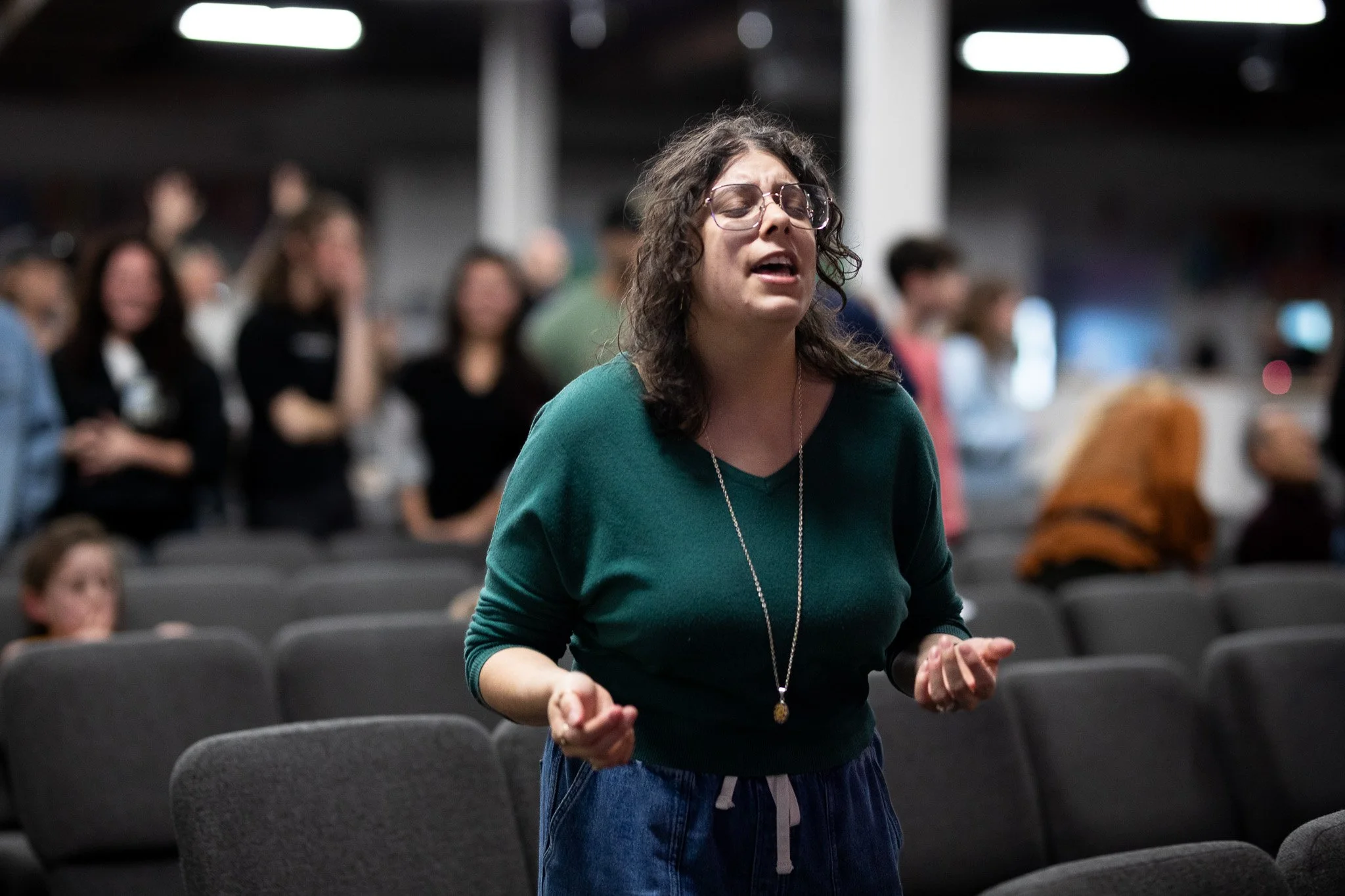 A woman with curly hair, glasses, wearing a green sweater and jeans, appears to be speaking or singing with her eyes closed in a crowded indoor space with rows of chairs and other people in the background.