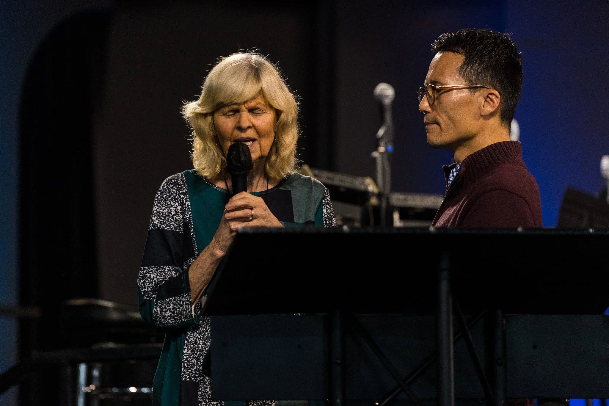 An elderly woman singing into a microphone and a young man standing beside her, both engaged in a performance indoors.