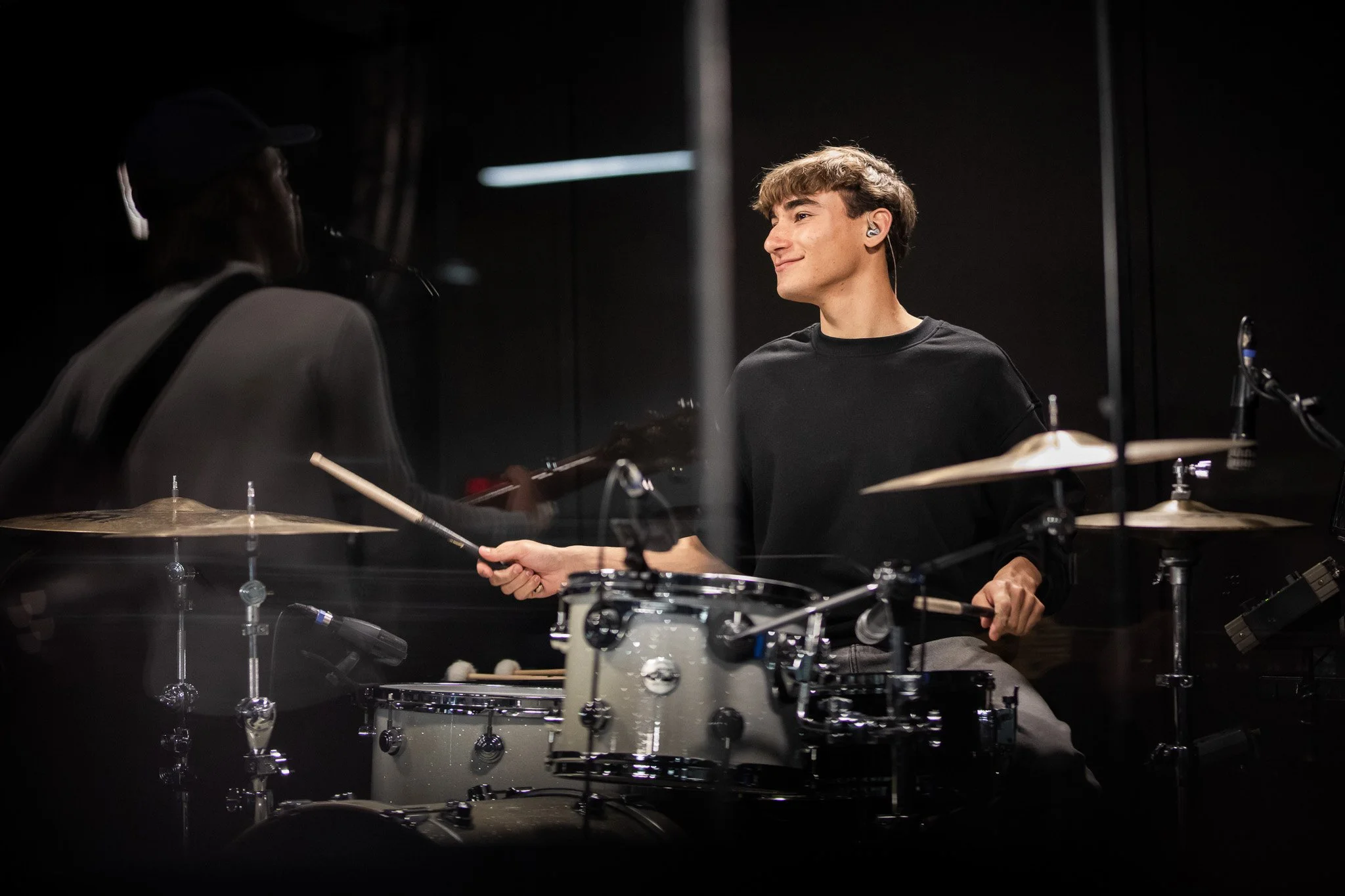 Young man playing drums in a dark studio, smiling, wearing a black shirt and in-ear monitors.