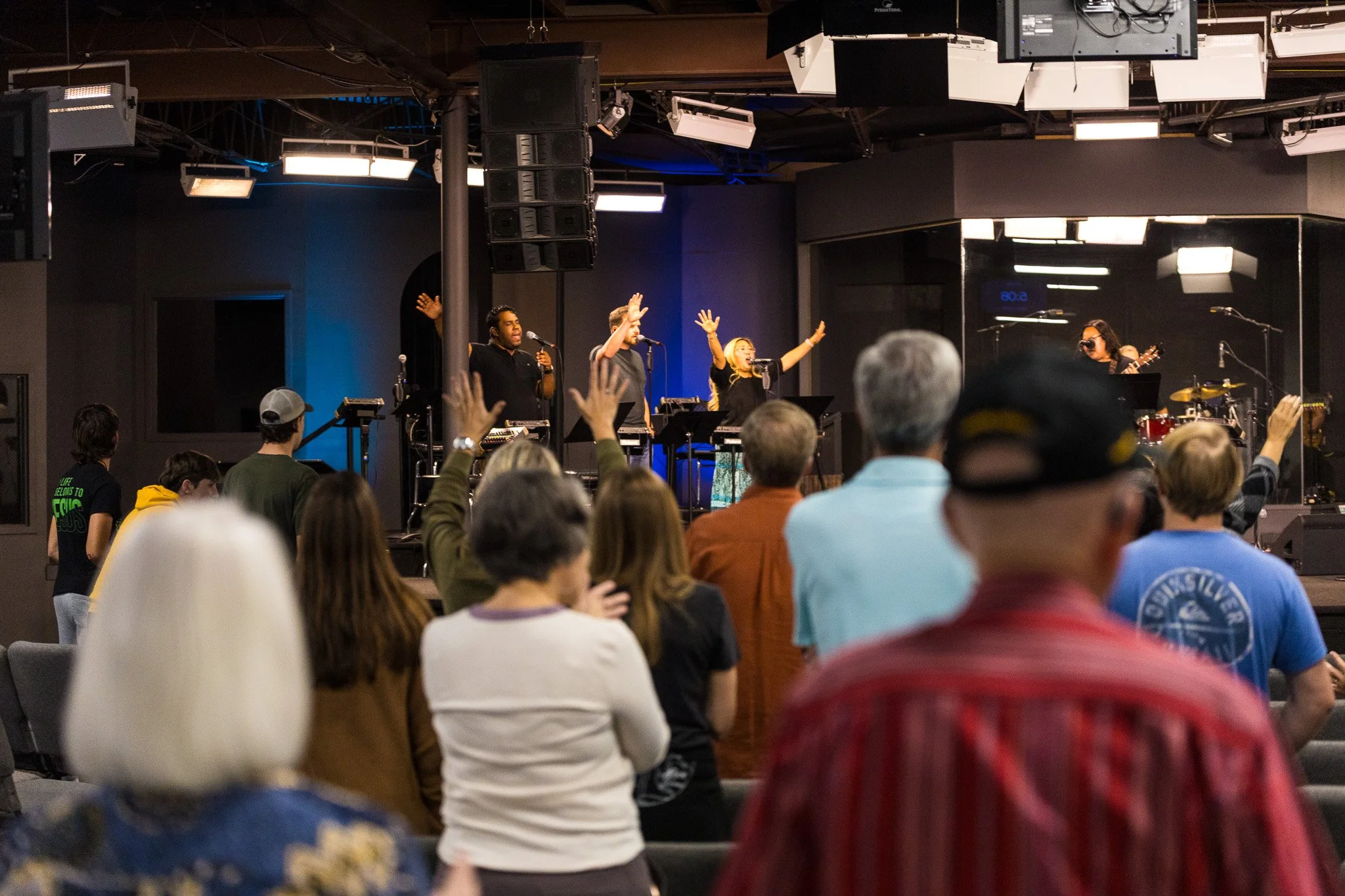 A live music performance on stage with four singers and a drummer, audience members raising their hands, in an indoor venue with professional lighting and sound equipment.