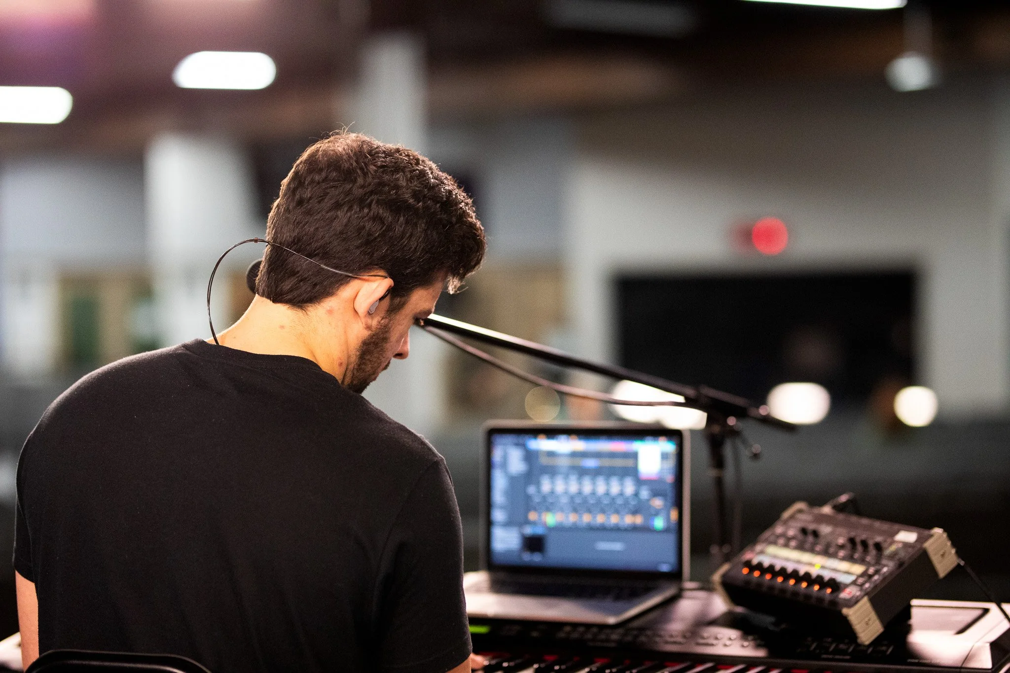 A man with dark hair and a beard wearing a black shirt, sitting at a music production desk, working with a laptop and various electronic music equipment, in a dimly lit room.