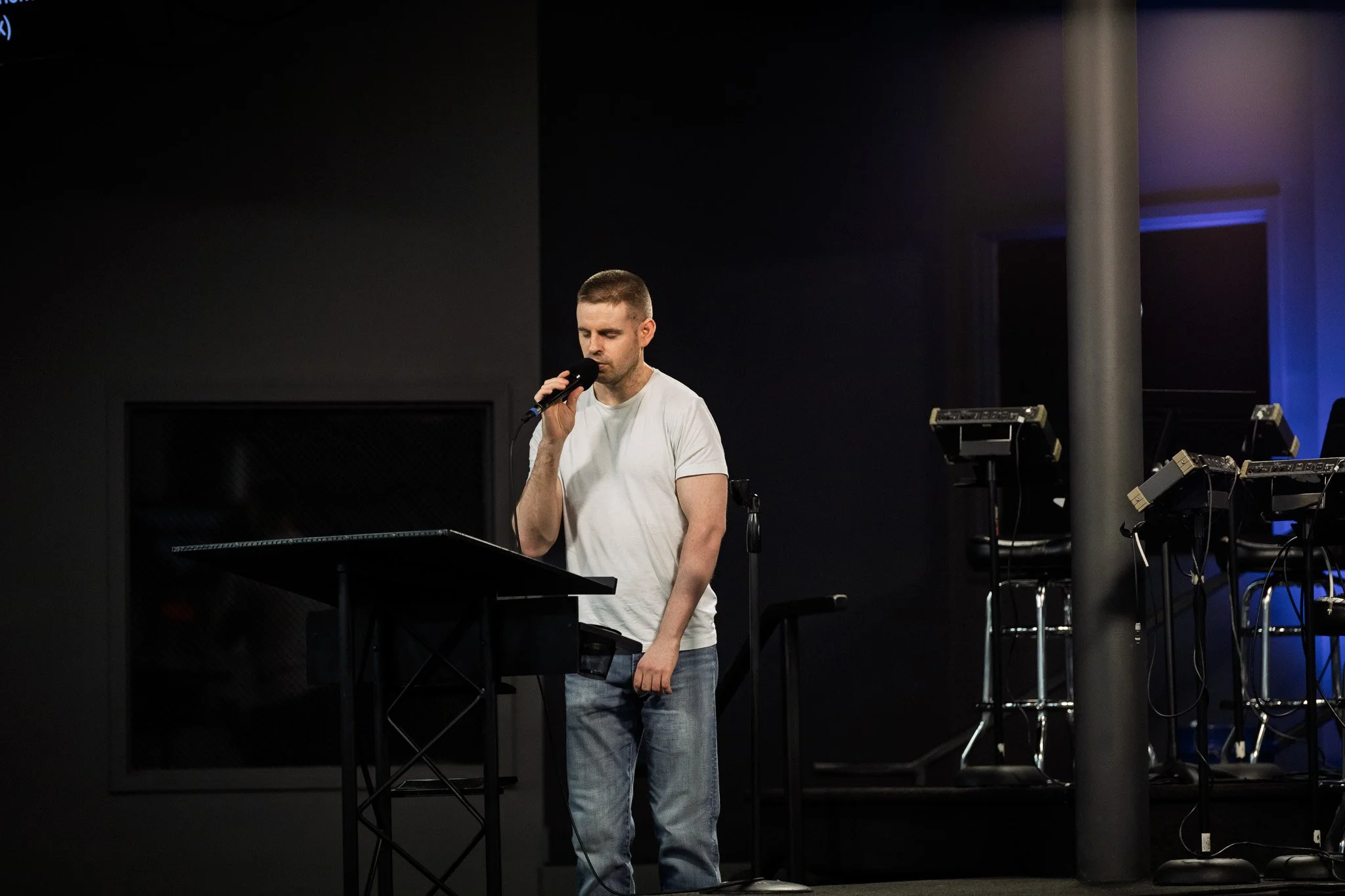 A young man with short hair and wearing a white t-shirt and jeans is standing at a black music stand, singing into a microphone in a dark indoor setting with blue-lit equipment around him.