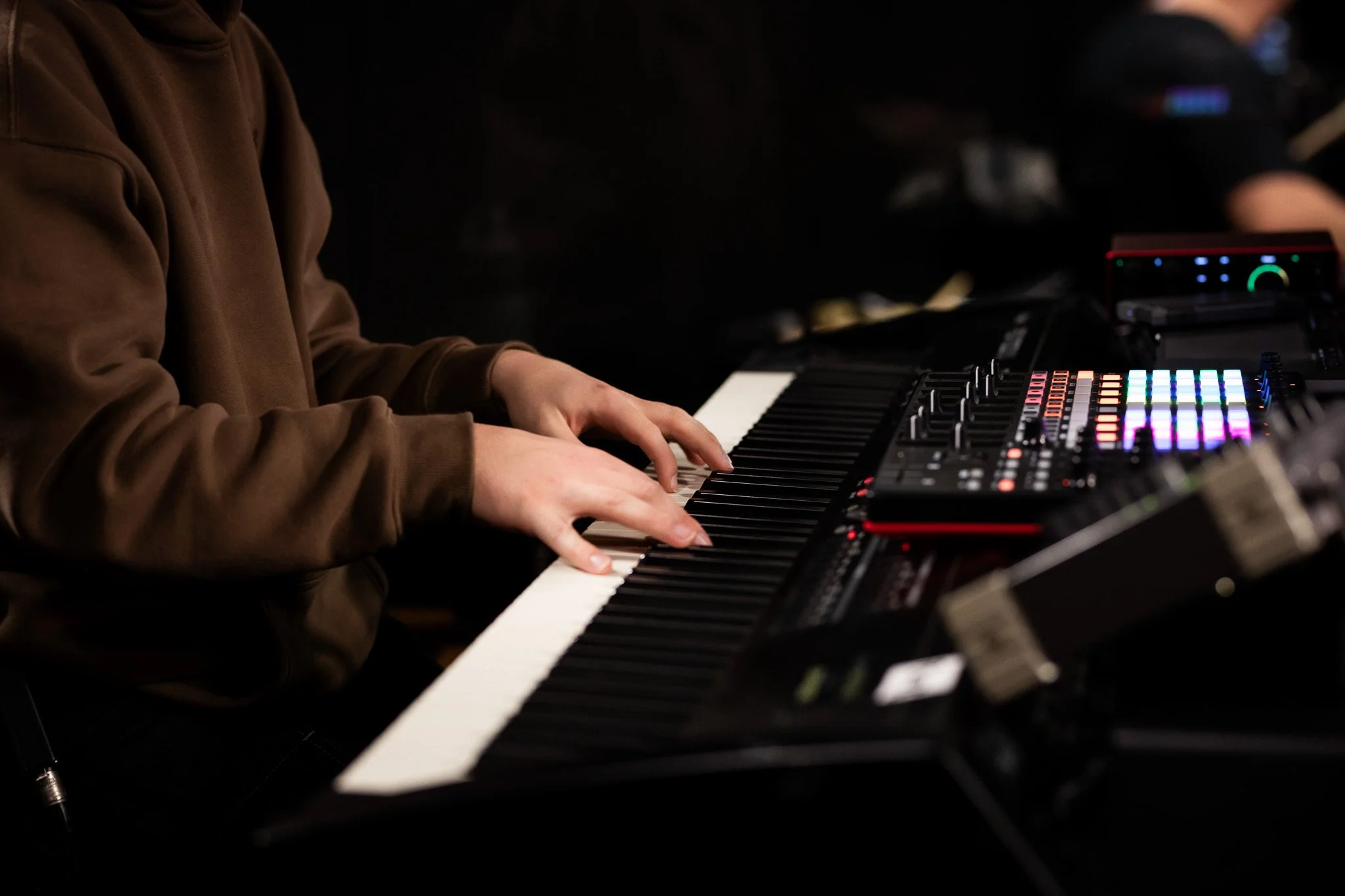 Person wearing a brown hoodie playing a keyboard with colorful lights in a dark environment.