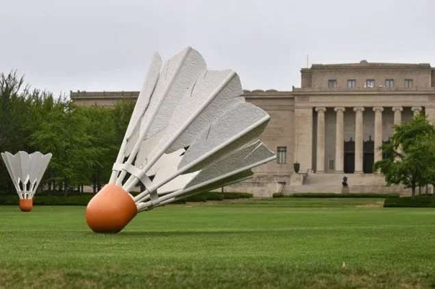 Large outdoor sculpture resembling a rocket with fins on a grassy area in front of a historic building.
