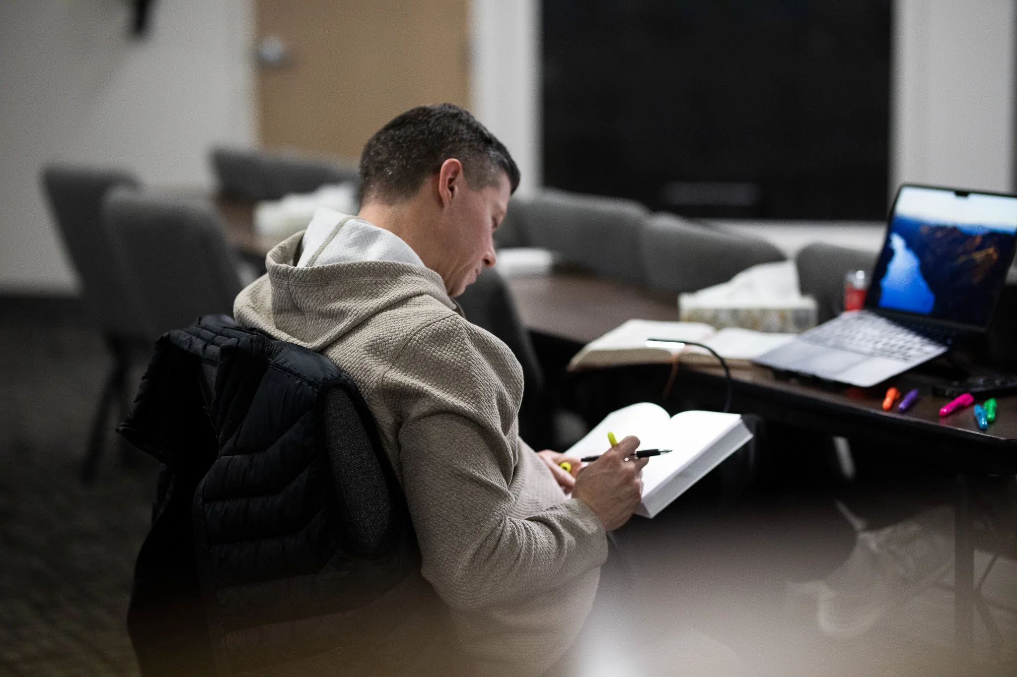 A young man with short dark hair is sitting at a table, reading or studying from an open book. He is wearing a beige hoodie and a black jacket. There's a laptop, some colorful pens, a tissue box, and a computer mouse on the table. The setting appears to be a classroom or study area with chairs and tables.