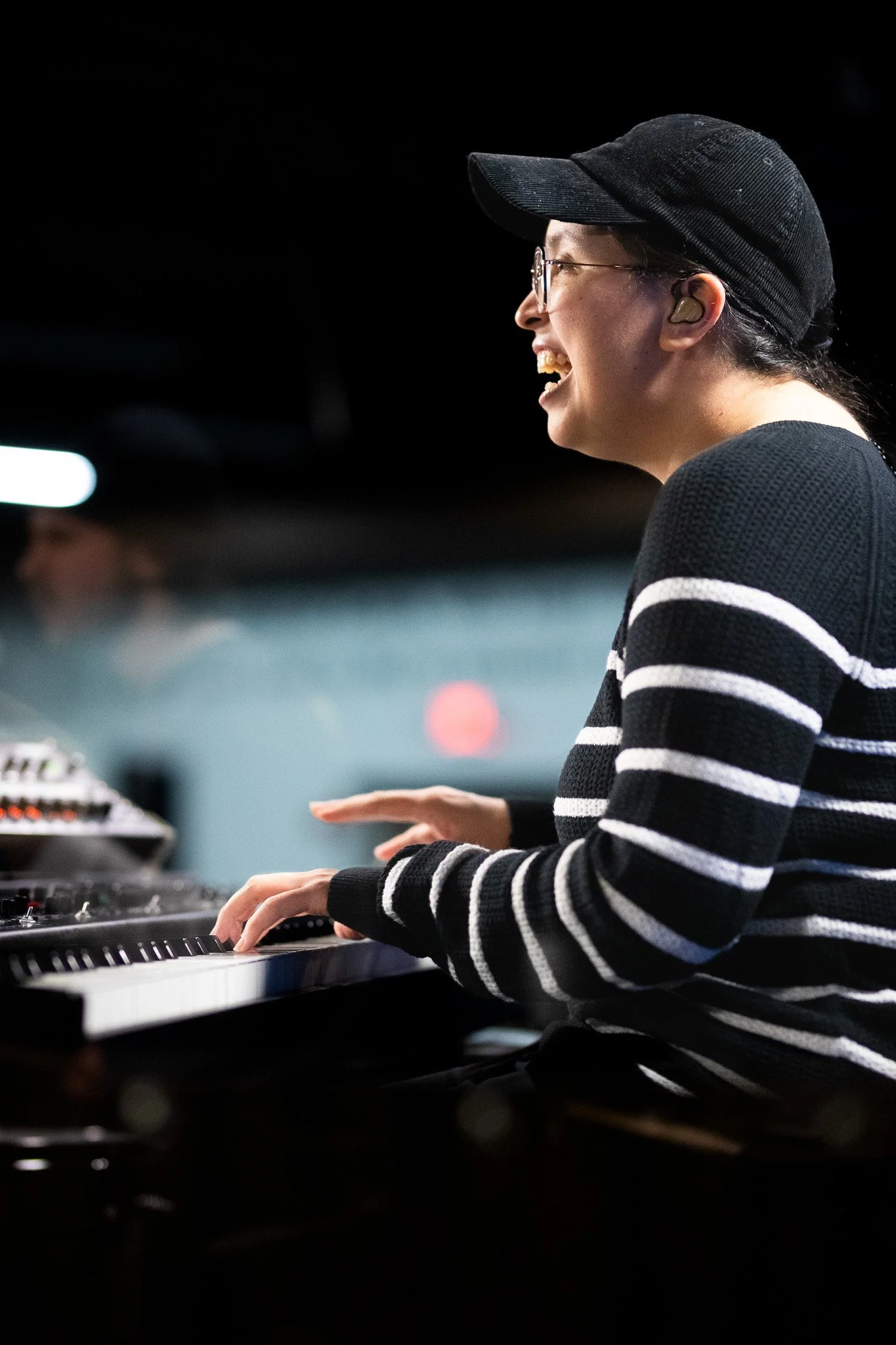 A person wearing glasses, a black cap, and a black and white striped sweater is playing a keyboard in a dimly lit setting, smiling and enjoying the music.