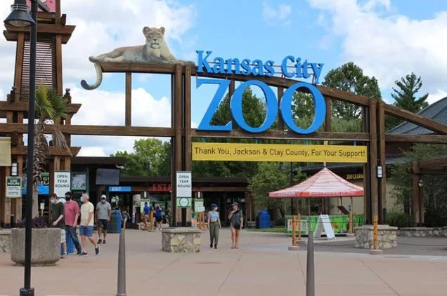 Entrance sign of Kansas City Zoo with a lion statue on top, visitors walking, and a red umbrella shelter.