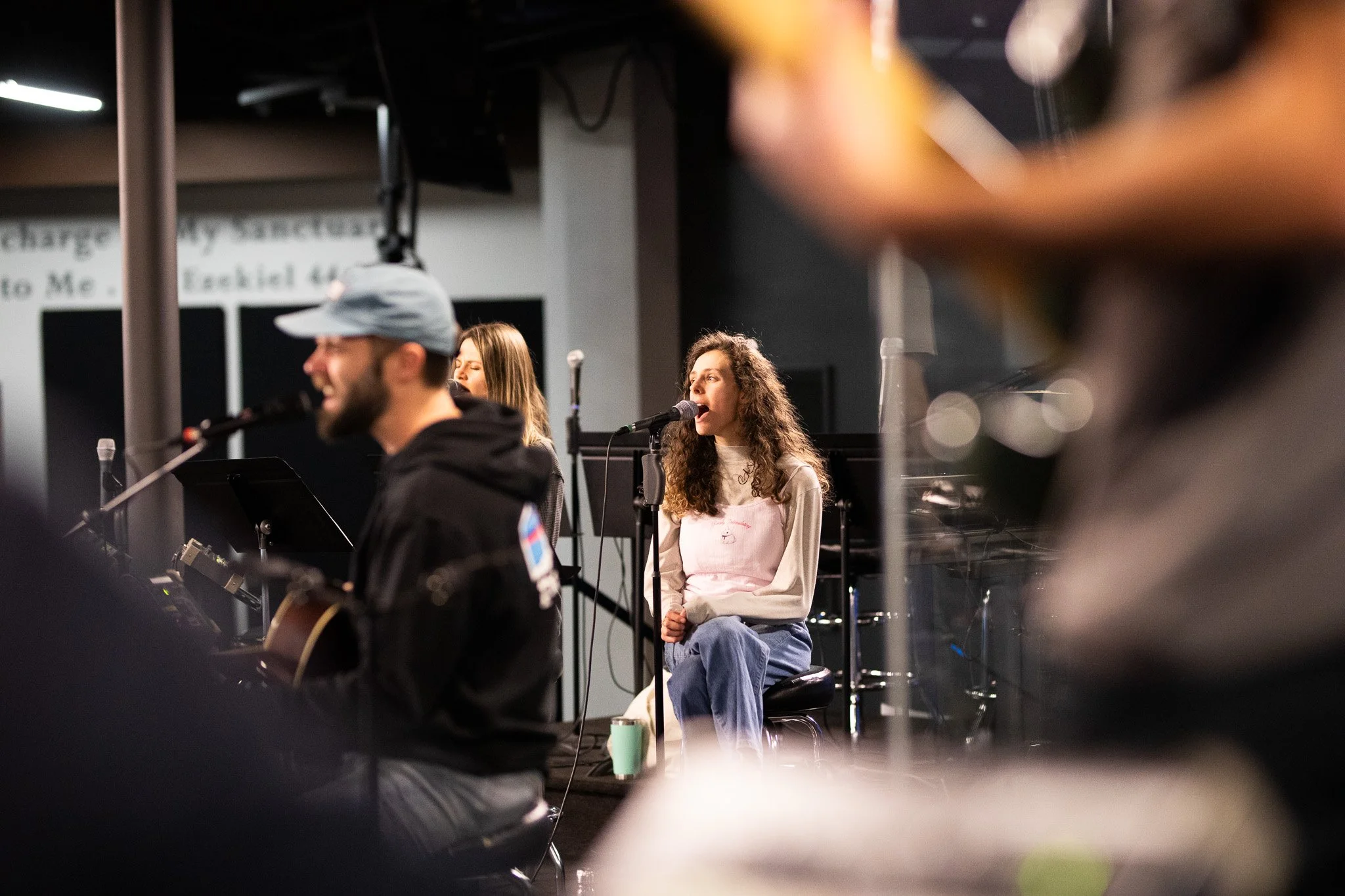 A woman with curly hair singing into a microphone, seated on a chair, with a man in a hat playing guitar in the foreground, in a dimly lit room during a live performance.