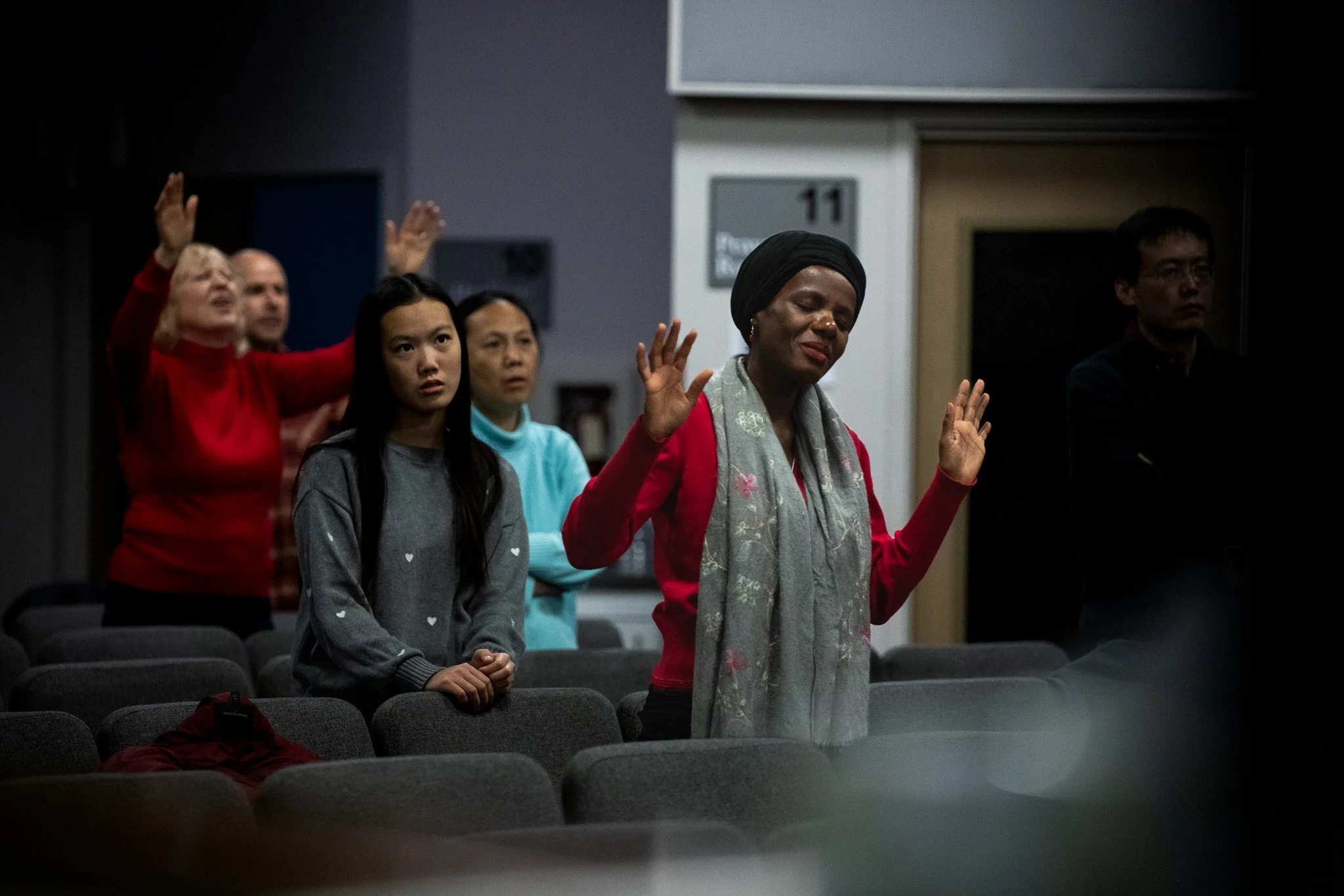 A group of people standing with eyes closed and hands raised in a prayer or meditation in a dark room.