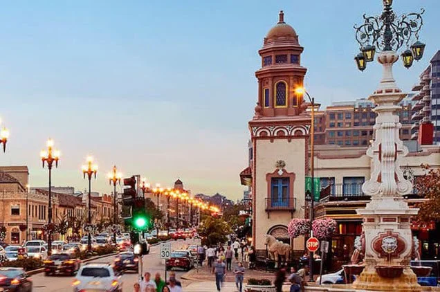 A lively city street at dusk with cars, pedestrians, ornate street lamps, and a historic pink and tan building with a clock tower, surrounded by modern buildings.