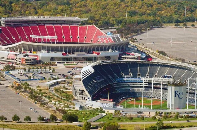 Aerial view of a large baseball stadium with red and black seats and surrounding parking lot, located in a green, wooded area.