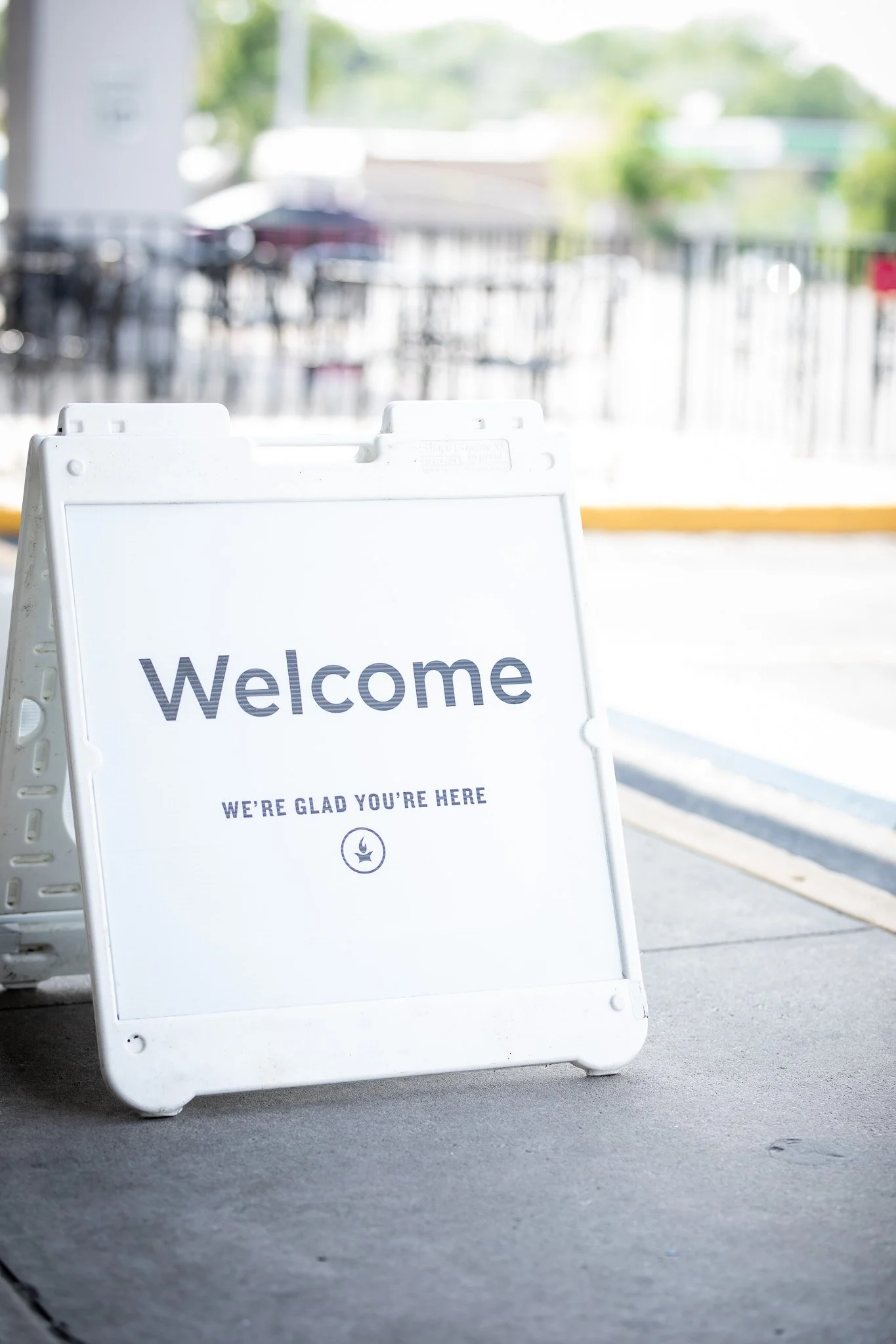 A white A-frame sign with the words 'Welcome' and 'We're glad you're here' printed on it, placed on a sidewalk outside a building.