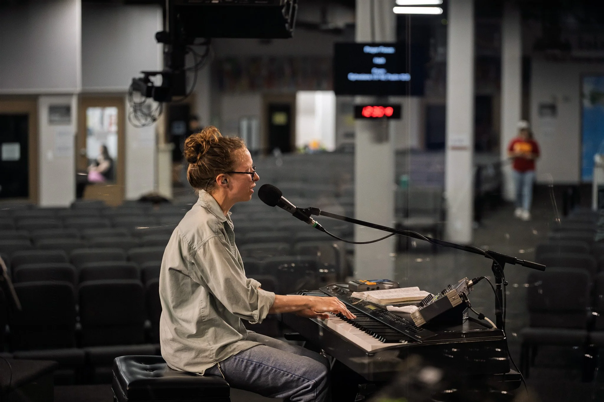 A woman with glasses and curly hair tied back, wearing a beige shirt, sits at a keyboard and sings into a microphone in a large, empty room with rows of chairs, some people in the background.