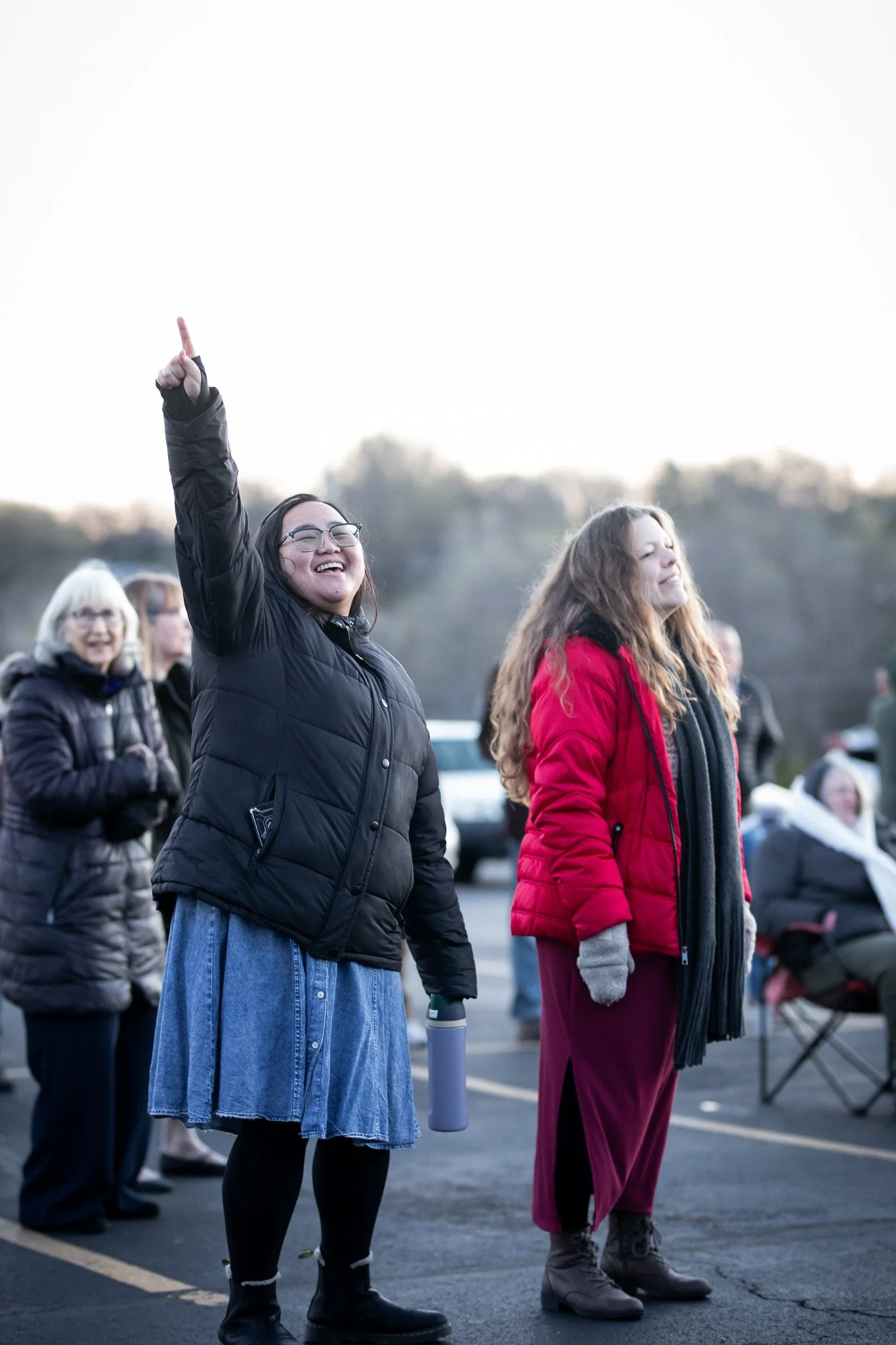 A group of people outdoors, some with closed eyes, standing on a parking lot during the daytime. One woman with glasses, wearing a black puffer jacket and a denim skirt, is smiling and raising her right hand with her index finger pointed up. Others are in jackets and warm clothing, appearing to participate in a gathering or event.