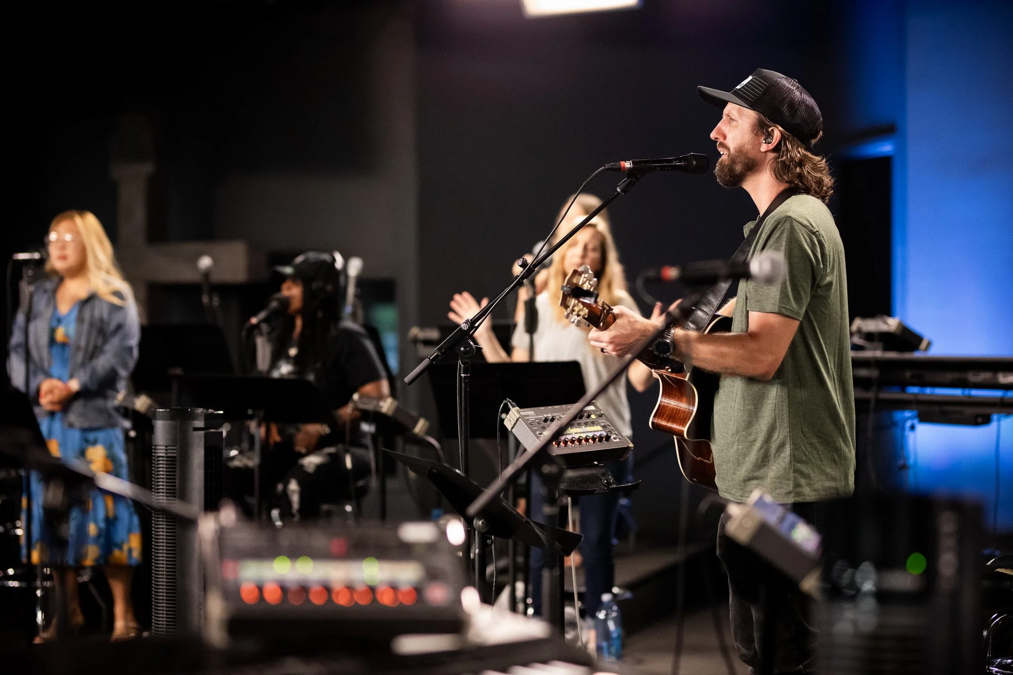 A man with long hair and a beard wearing a baseball cap is playing an acoustic guitar and singing into a microphone on stage. There are women in the background, some with microphones, likely part of a band or choir, in a dark room with stage lighting.