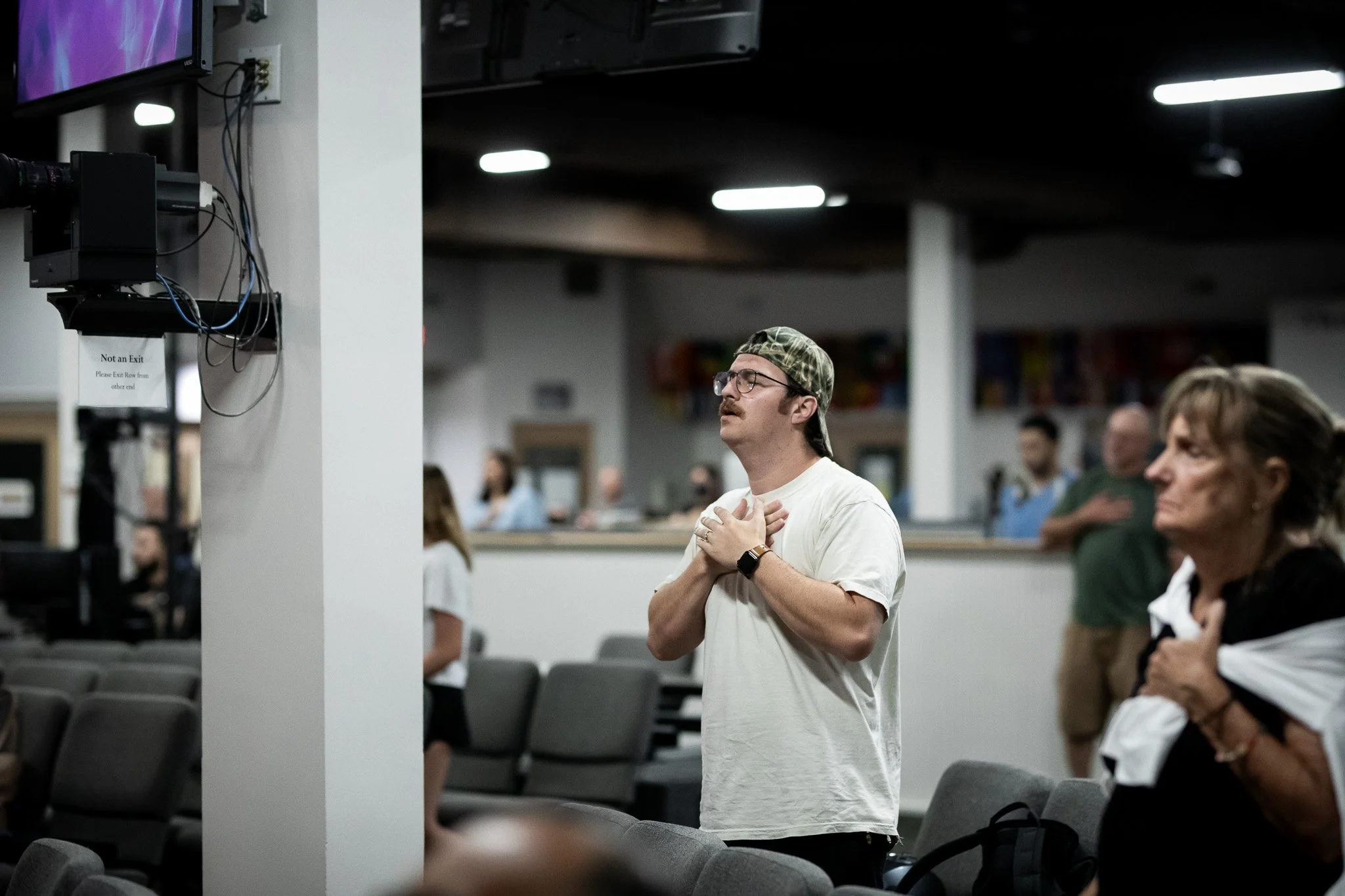 A man with glasses, a cap, and a white t-shirt standing with hands on chest in a room with people seated and some standing, under fluorescent lights.