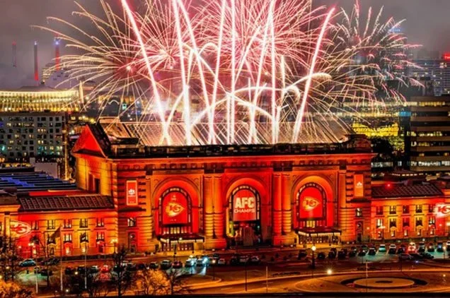 Fireworks display over a large, illuminated building with Kansas City Chiefs branding during a celebration.