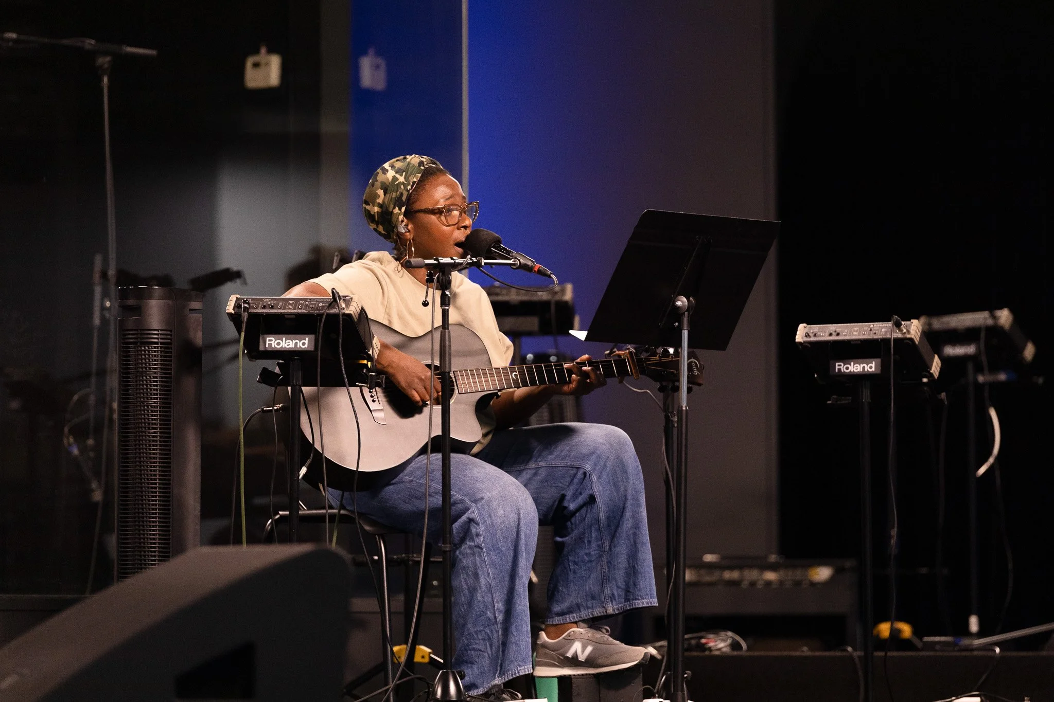 A woman wearing glasses and a camouflage headscarf is sitting on a stool, playing an acoustic guitar and singing into a microphone on a stage. She is surrounded by music equipment and a music stand.