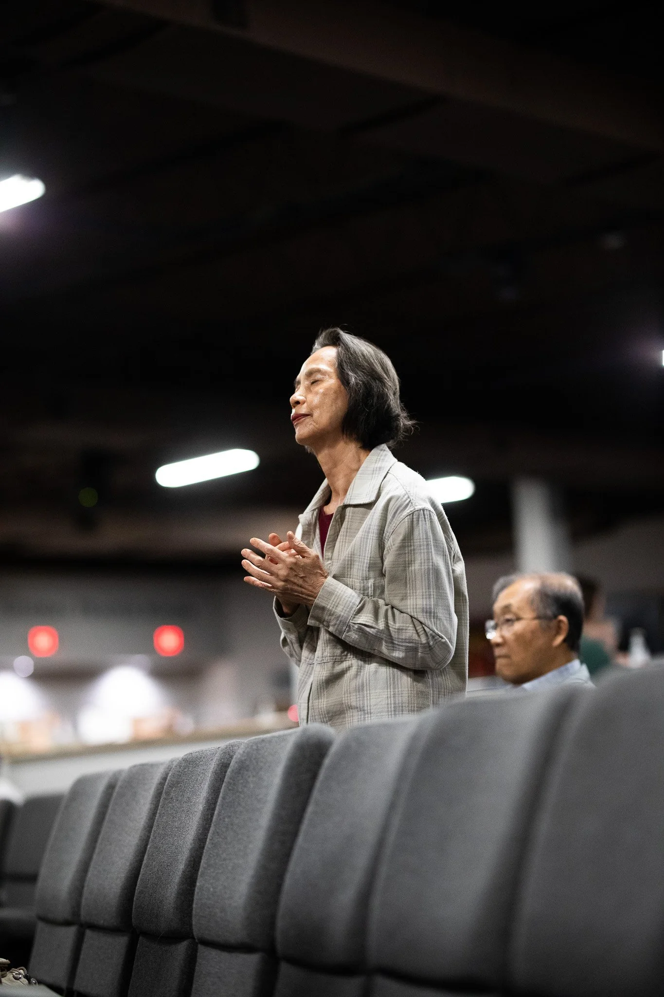 An elderly woman with short gray hair, wearing a light plaid shirt, standing with her eyes closed and hands clasped, in a dimly lit auditorium or conference room with empty gray chairs in the foreground and other seated people in the background.