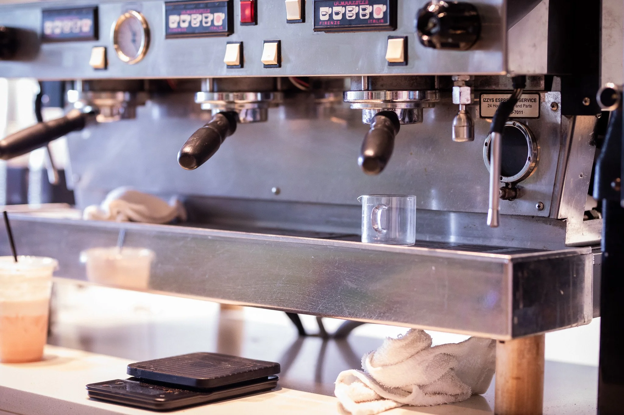 Close-up of a commercial espresso machine with coffee cups, a glass, and towels nearby, in a cafe setting.