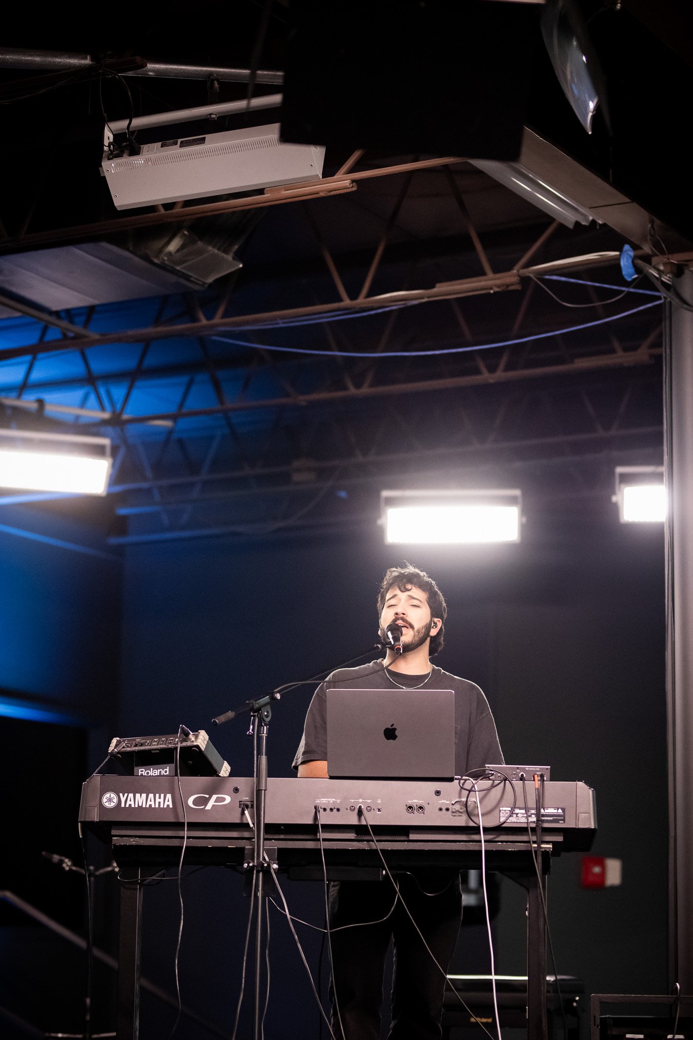 A man with dark curly hair and a beard is performing on stage, singing into a microphone, with a laptop and musical equipment in front of him. The stage has dark walls and a metal ceiling structure with bright stage lights overhead.