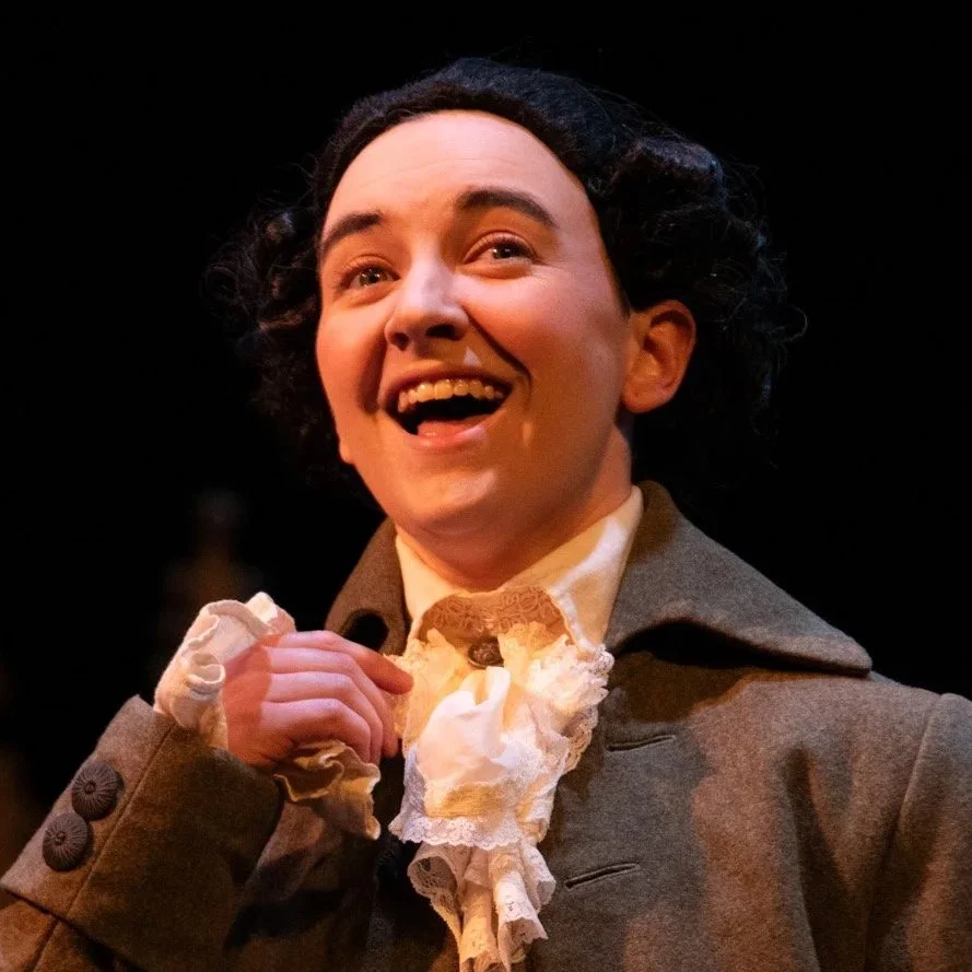 A woman dressed in historical clothing, smiling with an open mouth on a dark background.