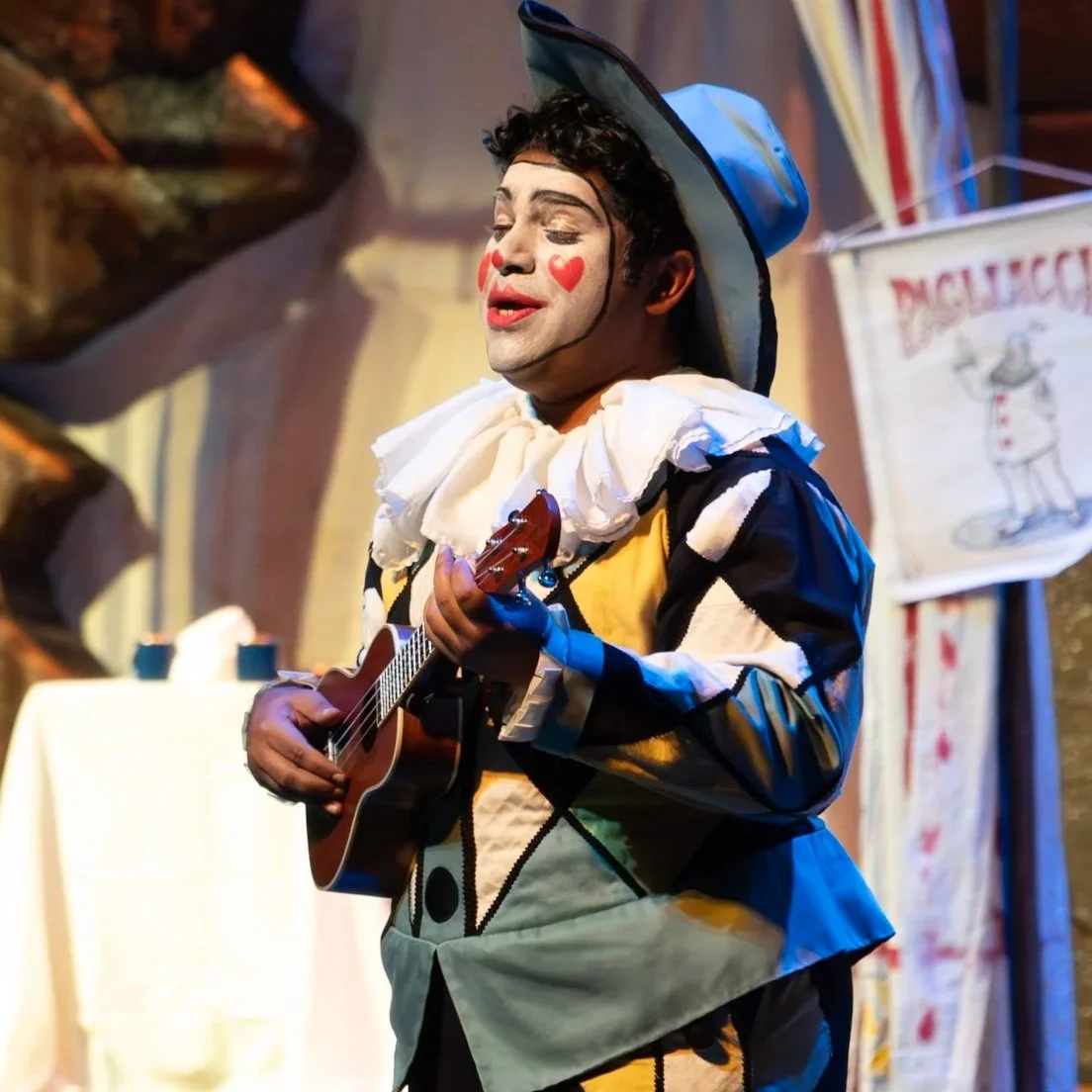 Andrew George as BEPPE with face paint, wearing a blue and yellow costume with a ruffled collar, playing a small guitar on stage.