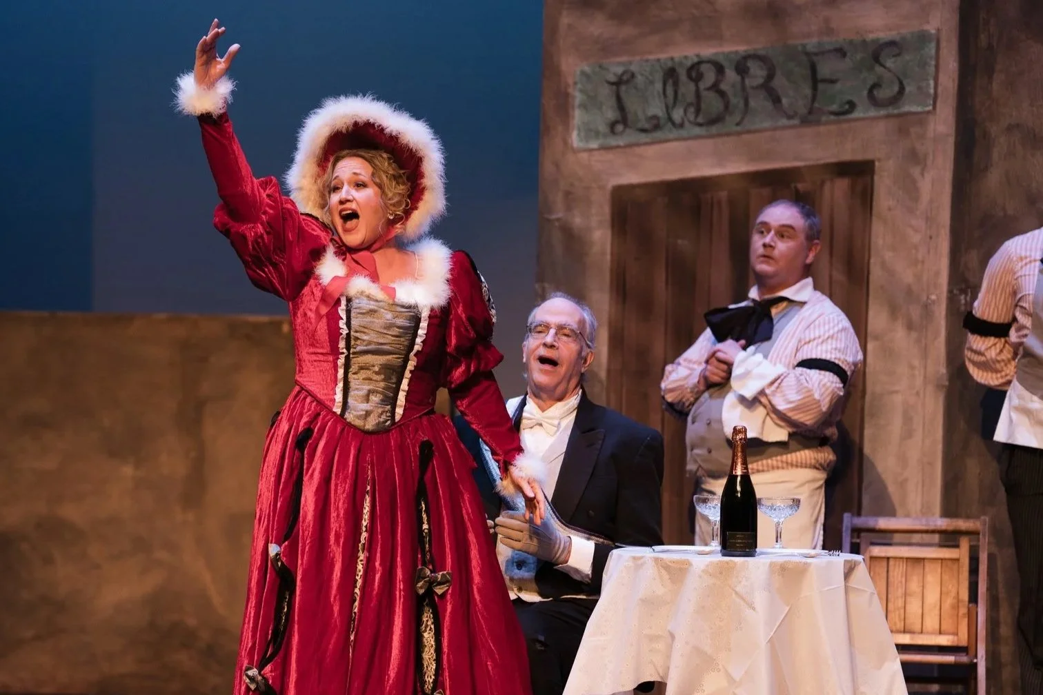 Julia Ebner in a red Victorian dress and matching hat gesturing passionately. Two men sit and stand nearby, one in a tuxedo and glasses, the other in a striped shirt holding a napkin, with a table holding a champagne bottle and glasses.