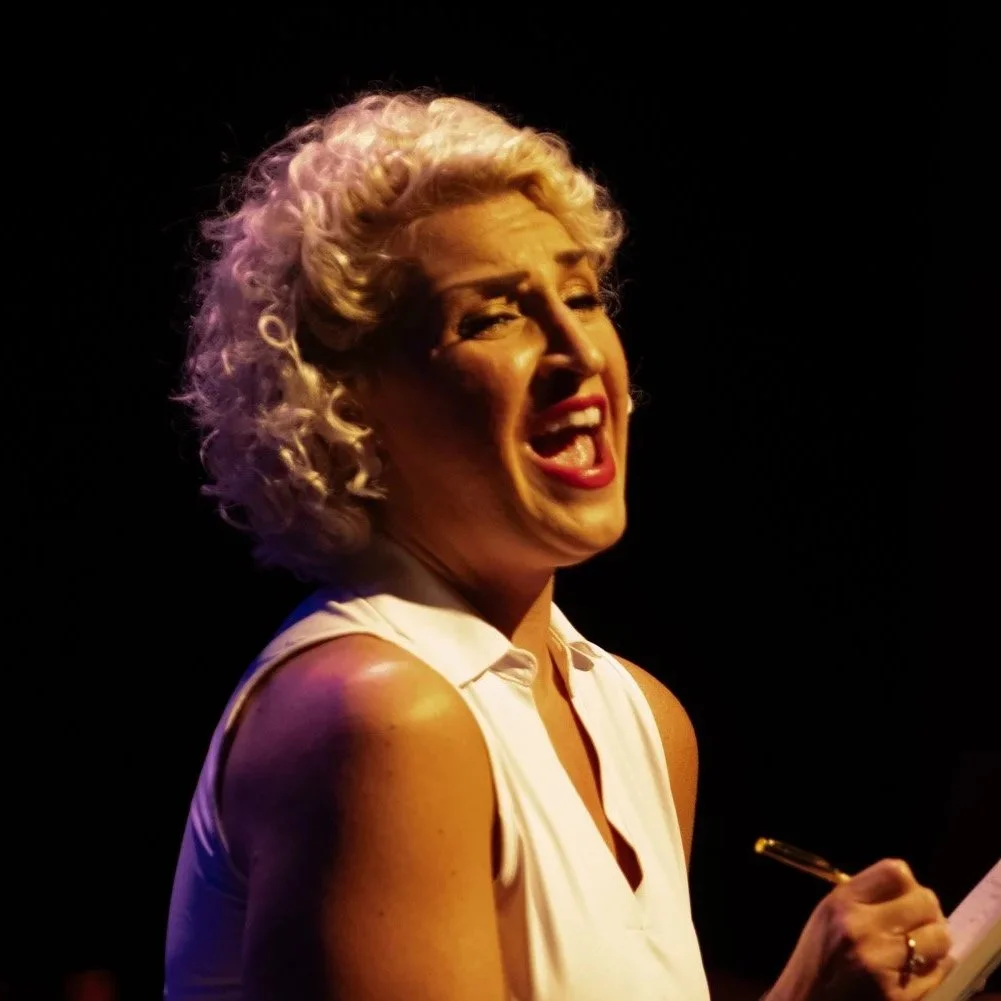 A woman with curly platinum blonde hair smiling and singing passionately on stage, holding a pen and paper, with a dark background.