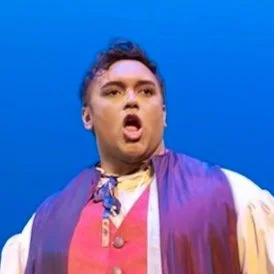 Person with curly hair and colorful clothing making an expressive face against a blue background.