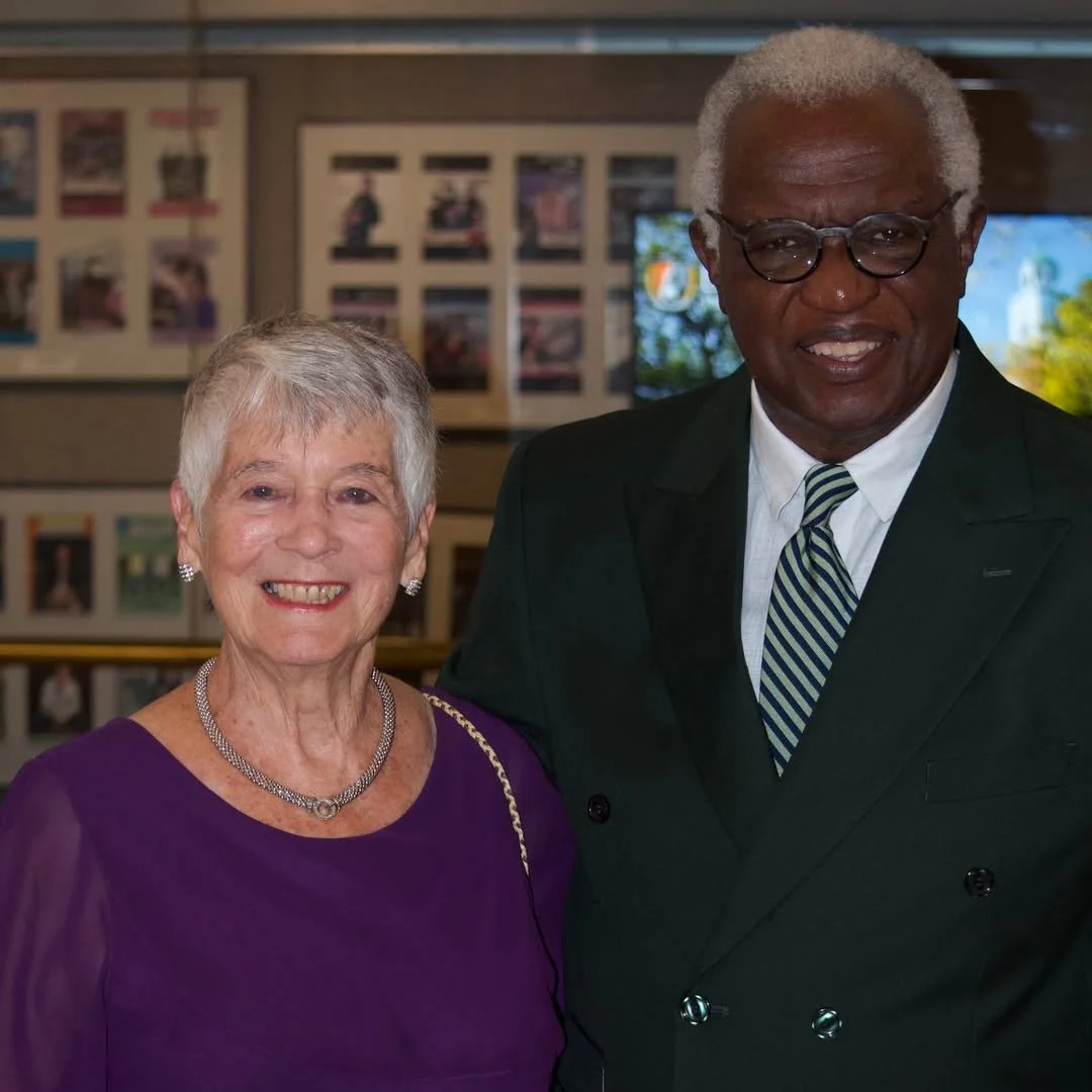 An elderly woman and an elderly man are standing together indoors, smiling at the camera. The woman is wearing a purple dress and a silver necklace, and the man is wearing a dark suit with a striped tie and glasses.
