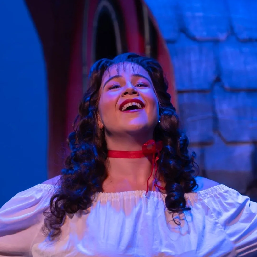 A woman with curly dark hair, wearing a white blouse and a red ribbon choker, smiling and looking upwards in bright theatrical lighting with a blue and brick background.