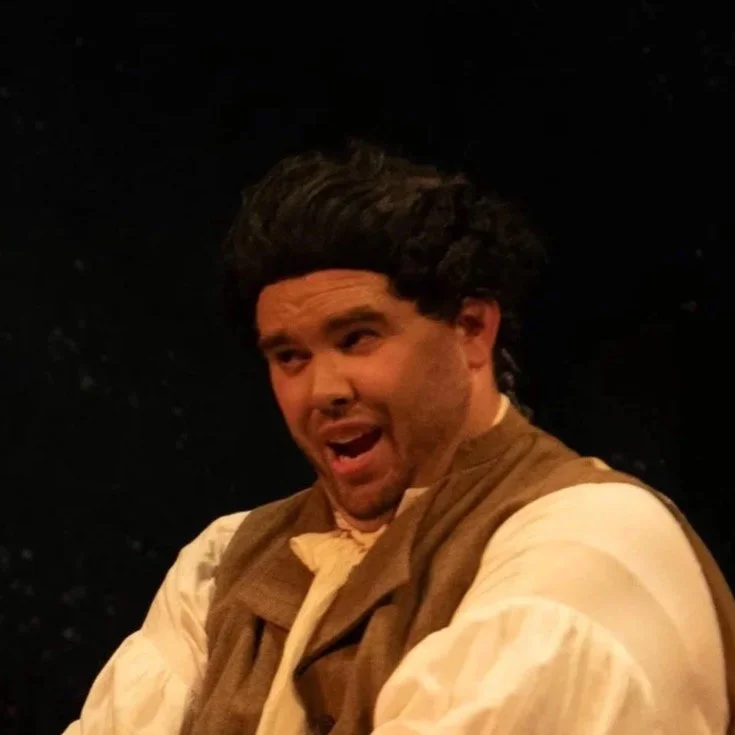 A man with dark, curly hair and a beard, wearing a beige shirt and brown vest, singing or speaking passionately on a dark stage.