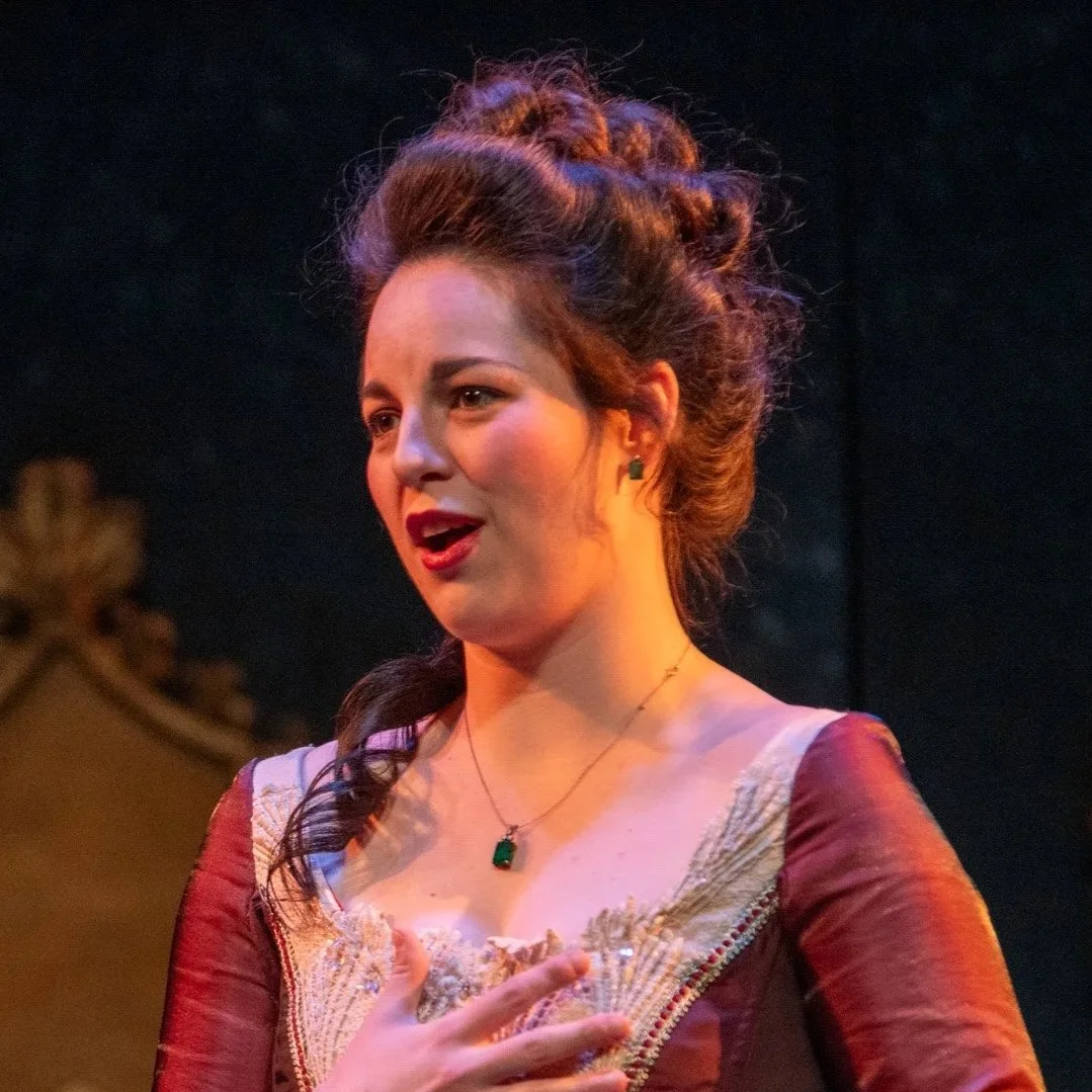 A woman with styled, reddish-brown hair, wearing a vintage dress with lace details, and a green gemstone necklace, performing in a theatrical play.