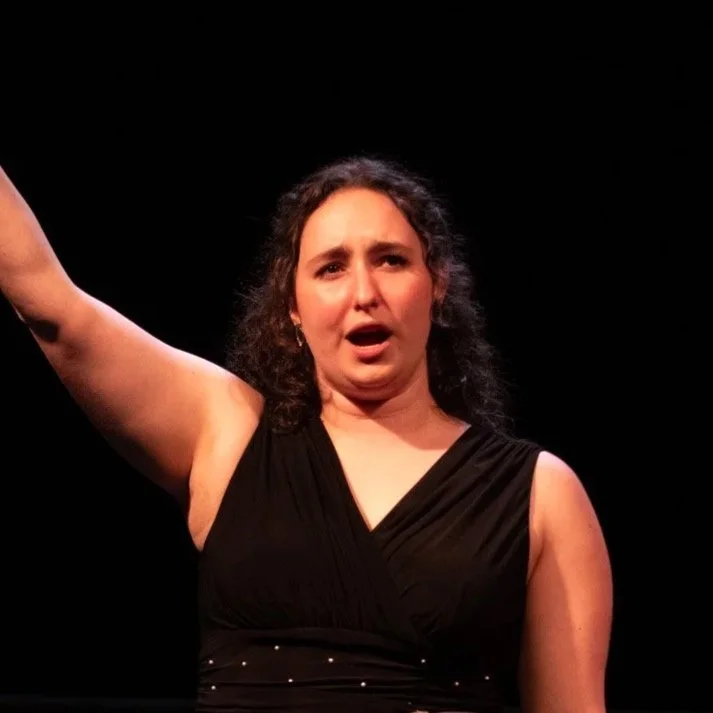 Woman with curly hair on stage, wearing a black sleeveless dress, raising her right arm, and speaking or singing, against a black background.