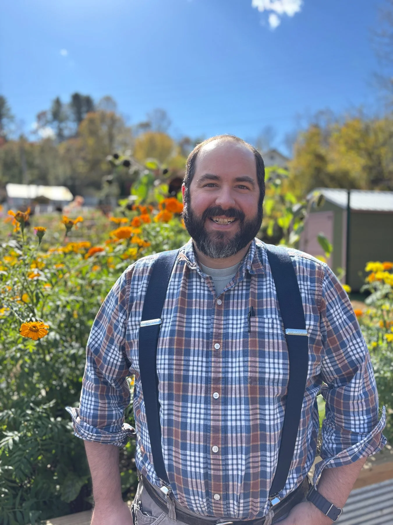 Smiling man with a beard and backpack standing in a garden filled with orange flowers on a sunny day.