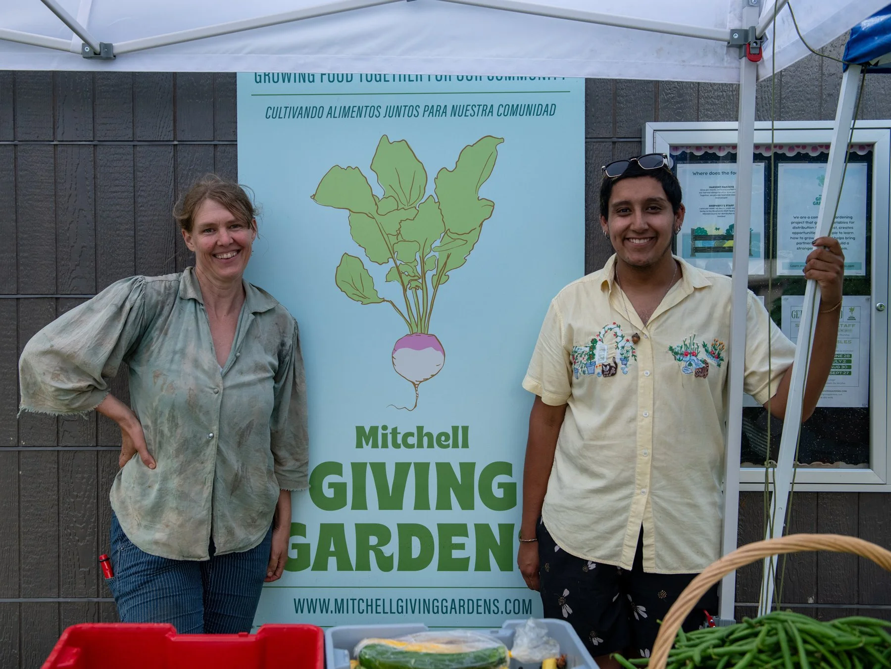 Two people standing in front of a Mitchell Giving Garden banner, smiling. The woman on the left is wearing a light-colored shirt with rolled-up sleeves and blue jeans, while the man on the right is wearing a yellow short-sleeved shirt with floral patterns and dark shorts, holding a tent pole.