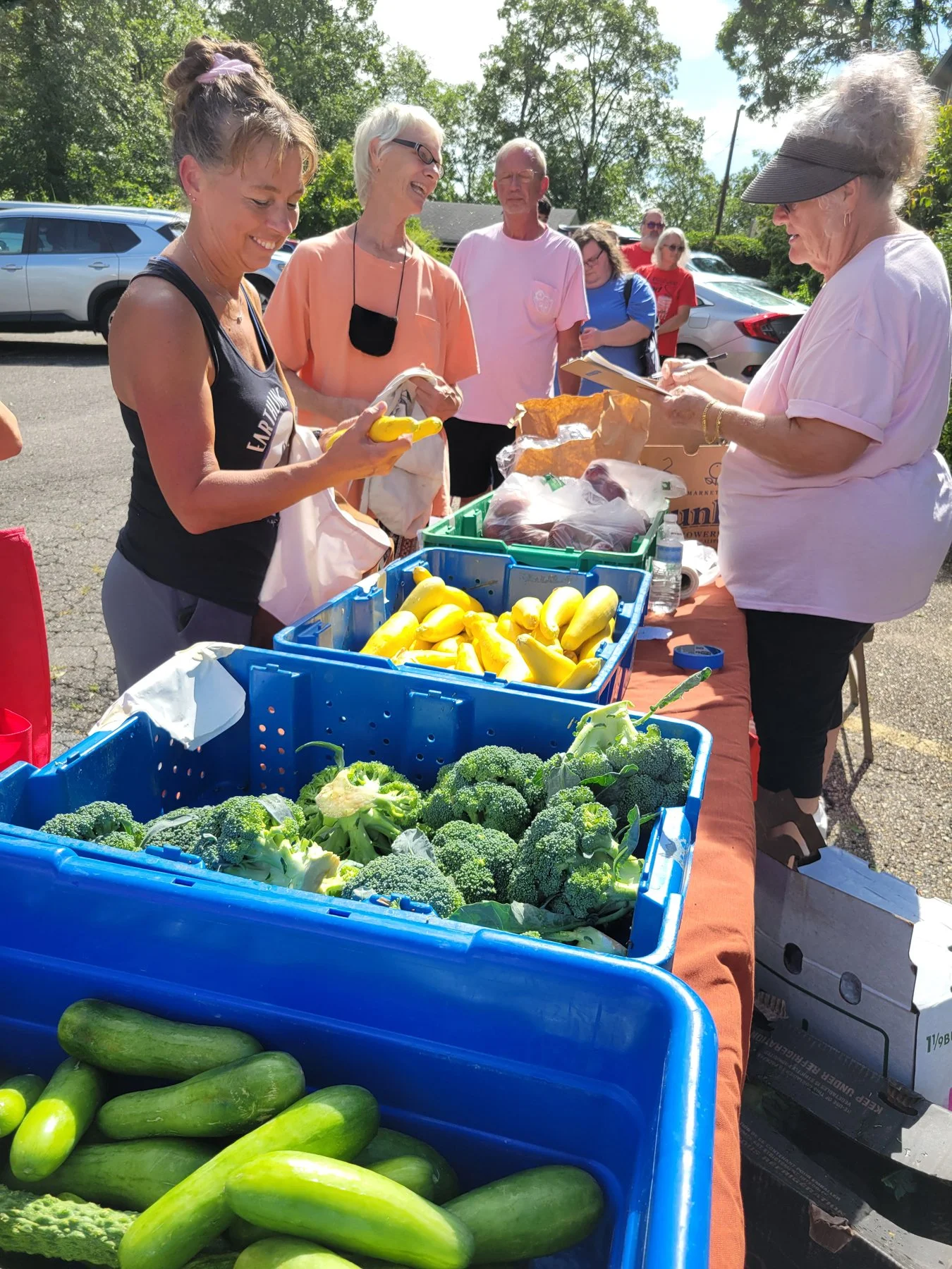 A group of people at an outdoor farmers market stand with fresh vegetables including zucchini, broccoli, and yellow squash, engaging in conversation and shopping on a sunny day.