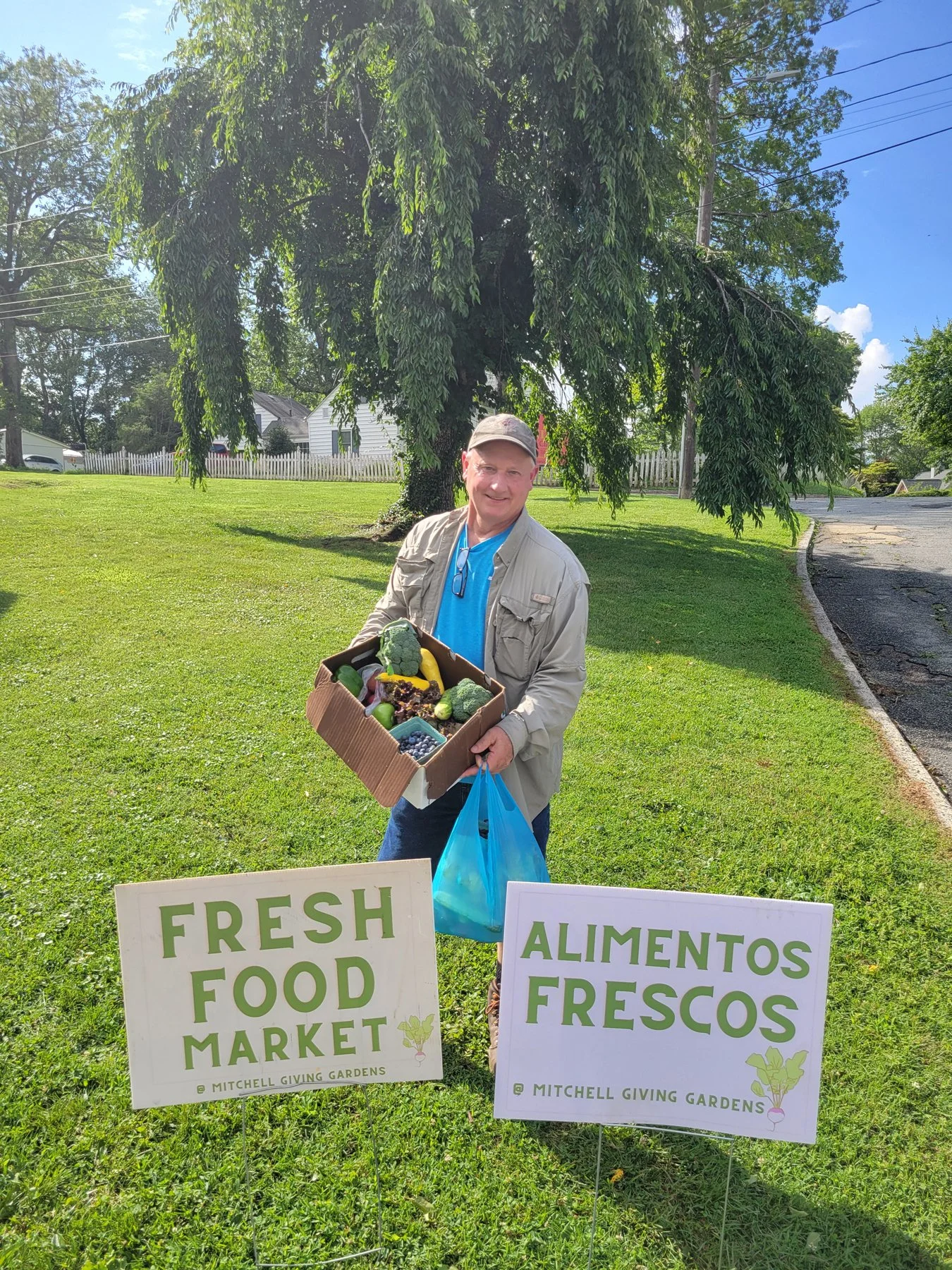 A man standing outdoors holding a box of fresh vegetables and fruits. There are two signs on the grass in front of him: one reads "FRESH FOOD MARKET" and the other "ALIMENTOS FRESCOS." The background features a large tree, a grassy area, a white fence, and a road with power lines overhead.