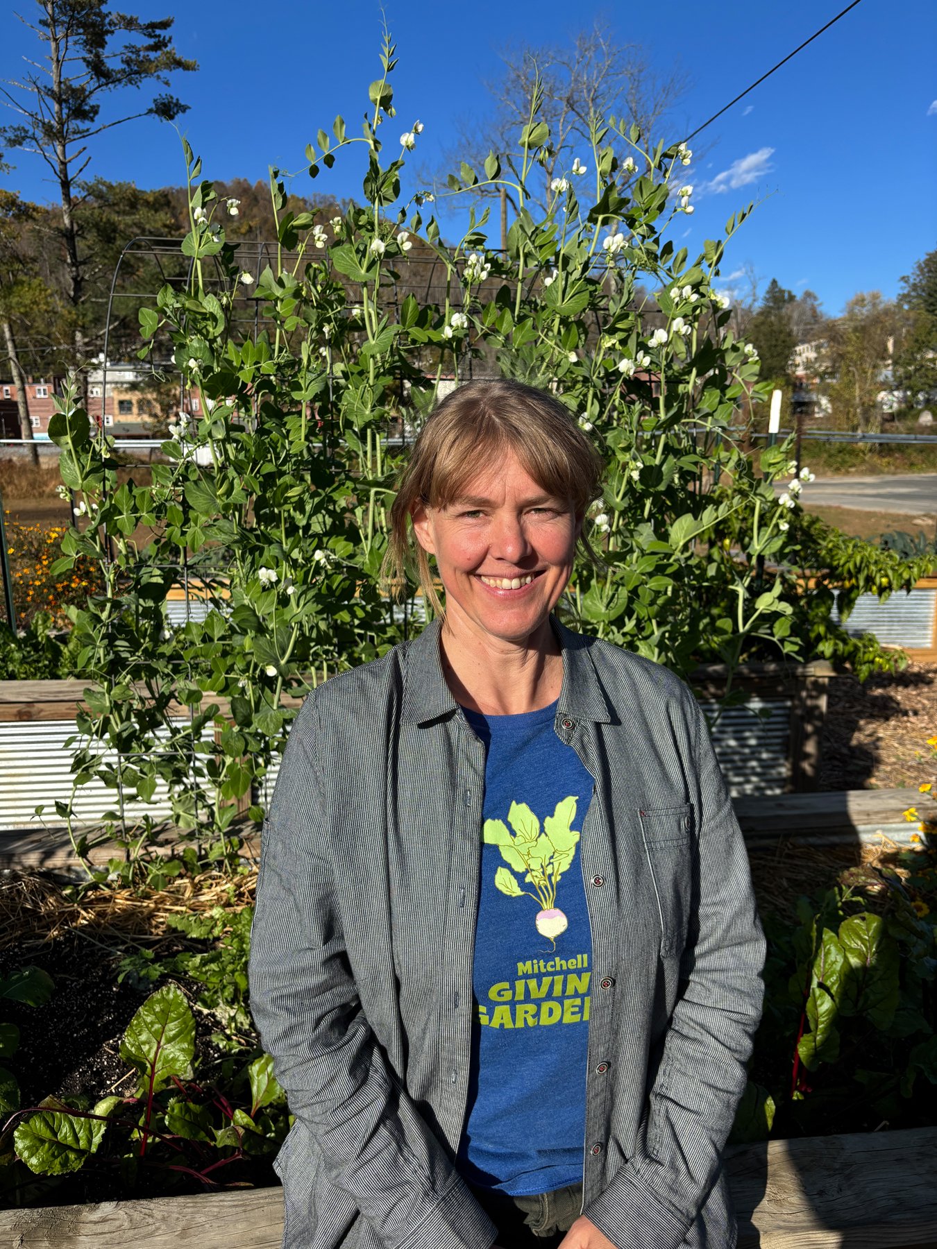 A woman standing in a garden, smiling, with tall green bean plants behind her, wearing a Mitchell Givin Garden t-shirt and a gray button-up shirt.