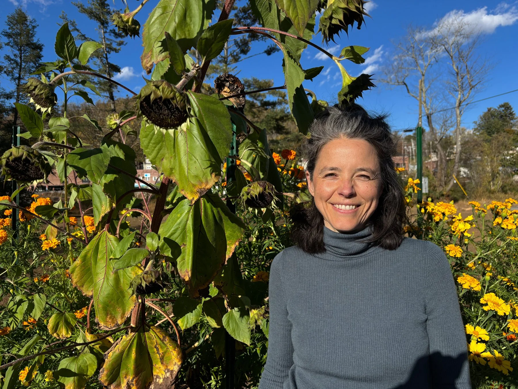 A woman with dark brown and grey peppered hair in a ponytail, smiling outdoors, standing among yellow and maroon flowers with a sunflower in the background, under a blue sky with some clouds.