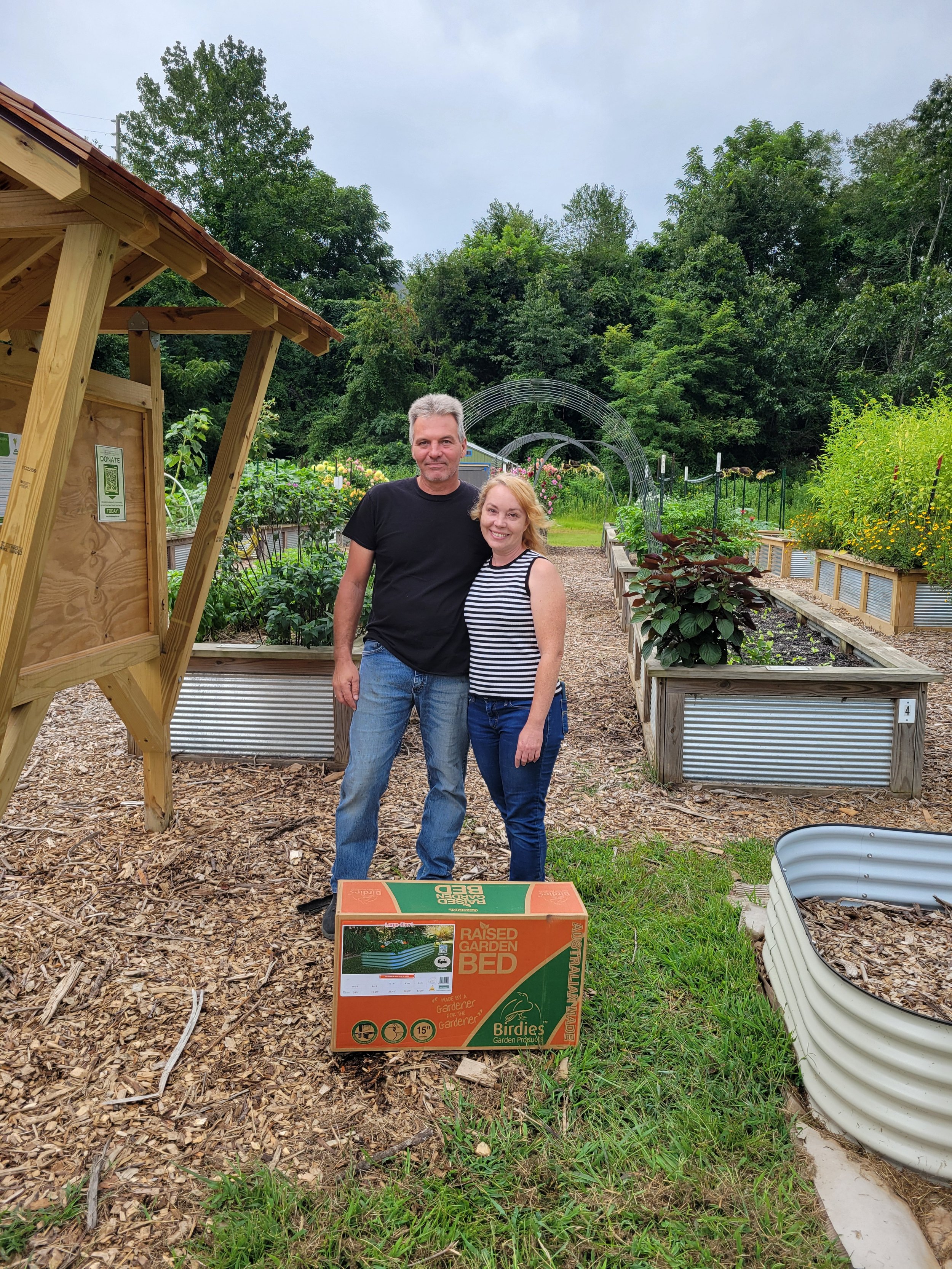 Two people standing in the mitchell giving garden with raised beds, holding a box labeled "Raised Garden Bed." They are outdoors with trees and a cloudy sky in the background.