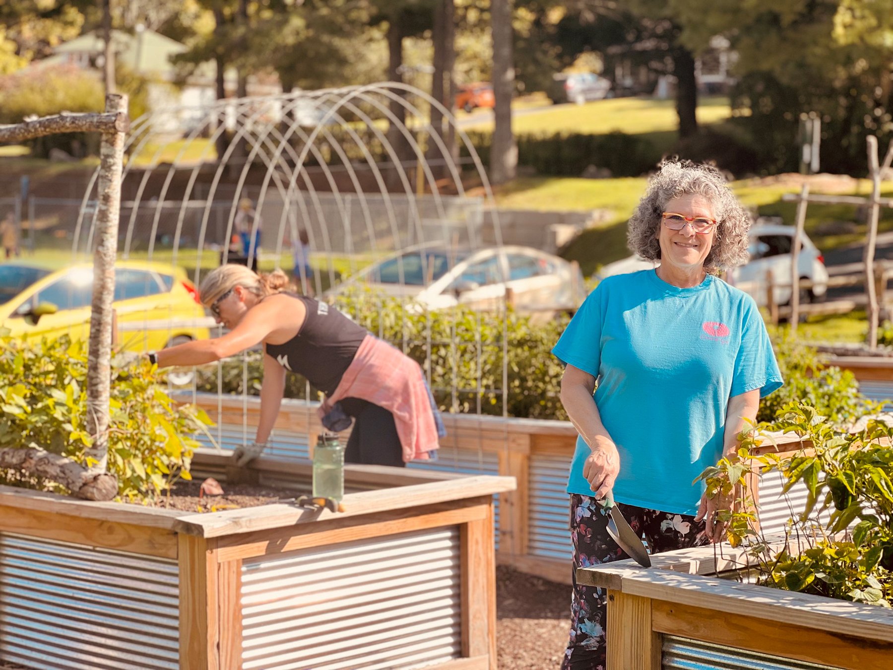 Smiling woman with curly gray hair and glasses, wearing a blue T-shirt and floral pants, gardening in the mitchell giving garden's community garden with raised wooden beds and other volunteers in the background.